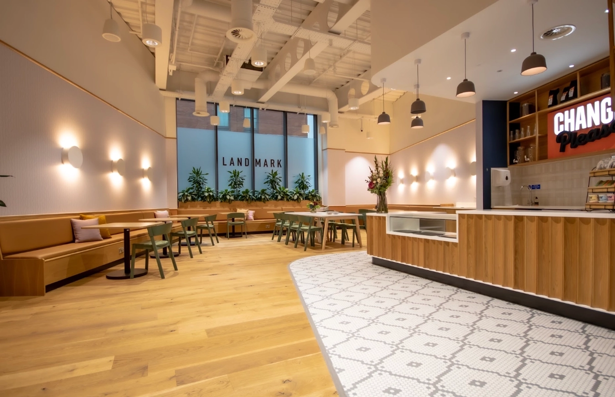 Modern cafe interior with wooden floors, tiled counter area, tan seating, green chairs, wall lights, and large windows with "LANDMARK" visible outside—perfect for a casual workspace or informal meeting room setting.