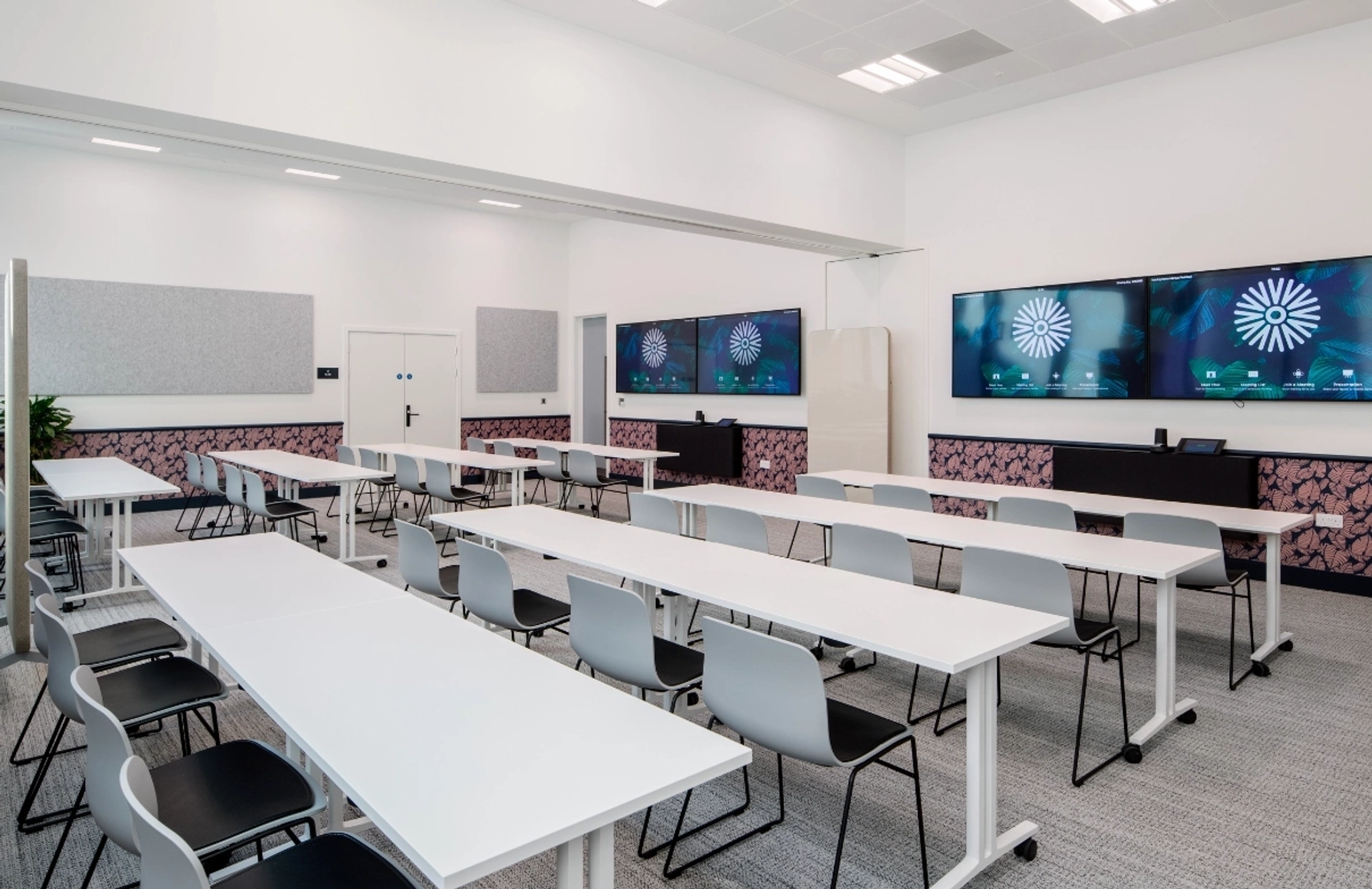 Modern classroom with white tables and gray chairs arranged in rows, resembling a coworking space in Manchester, featuring three large wall-mounted screens at the front displaying a loading symbol.