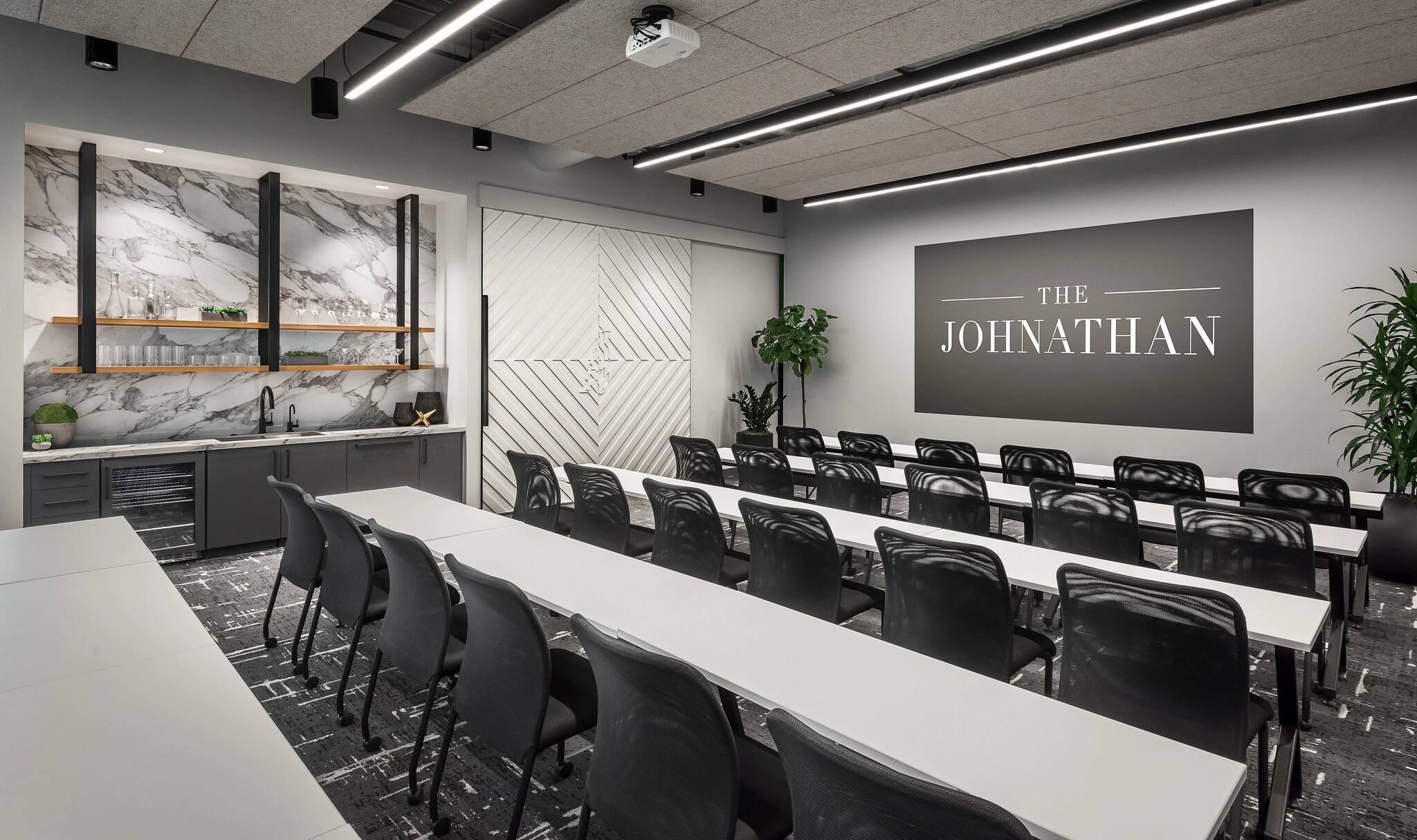 Modern conference room with rows of black chairs and white tables facing a wall with "The Johnathan" sign, perfect for coworking. A marble-patterned kitchenette is on the left, and plants decorate the space, creating an inviting office atmosphere.