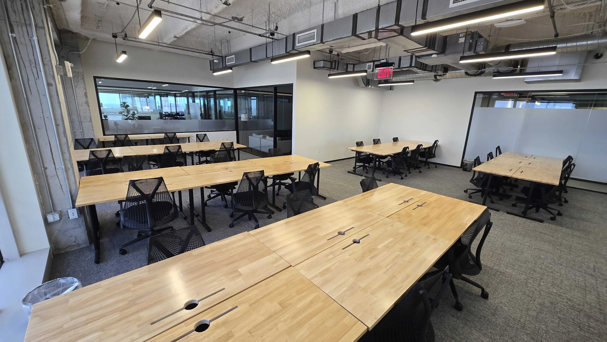 An empty modern coworking space with wooden desks and black mesh chairs arranged in rows. Large windows and glass partition walls are visible in the background, creating a bright and open workspace.