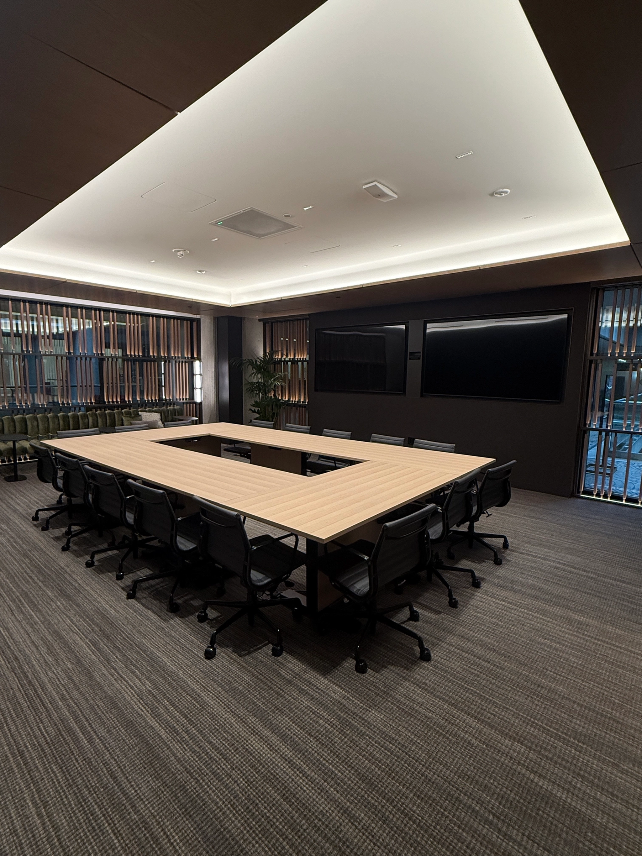 A modern meeting room with a large U-shaped table and black chairs, this versatile workspace features a wall-mounted screen and floor-to-ceiling windows with blinds.