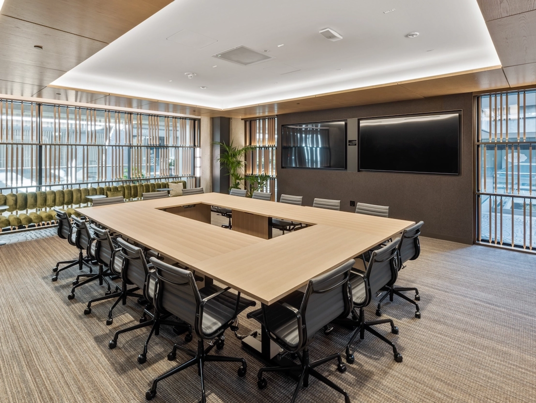 Modern coworking conference room with a large U-shaped wooden table, several office chairs, two large wall-mounted screens, and floor-to-ceiling windows with blinds—perfect for any collaborative workspace.