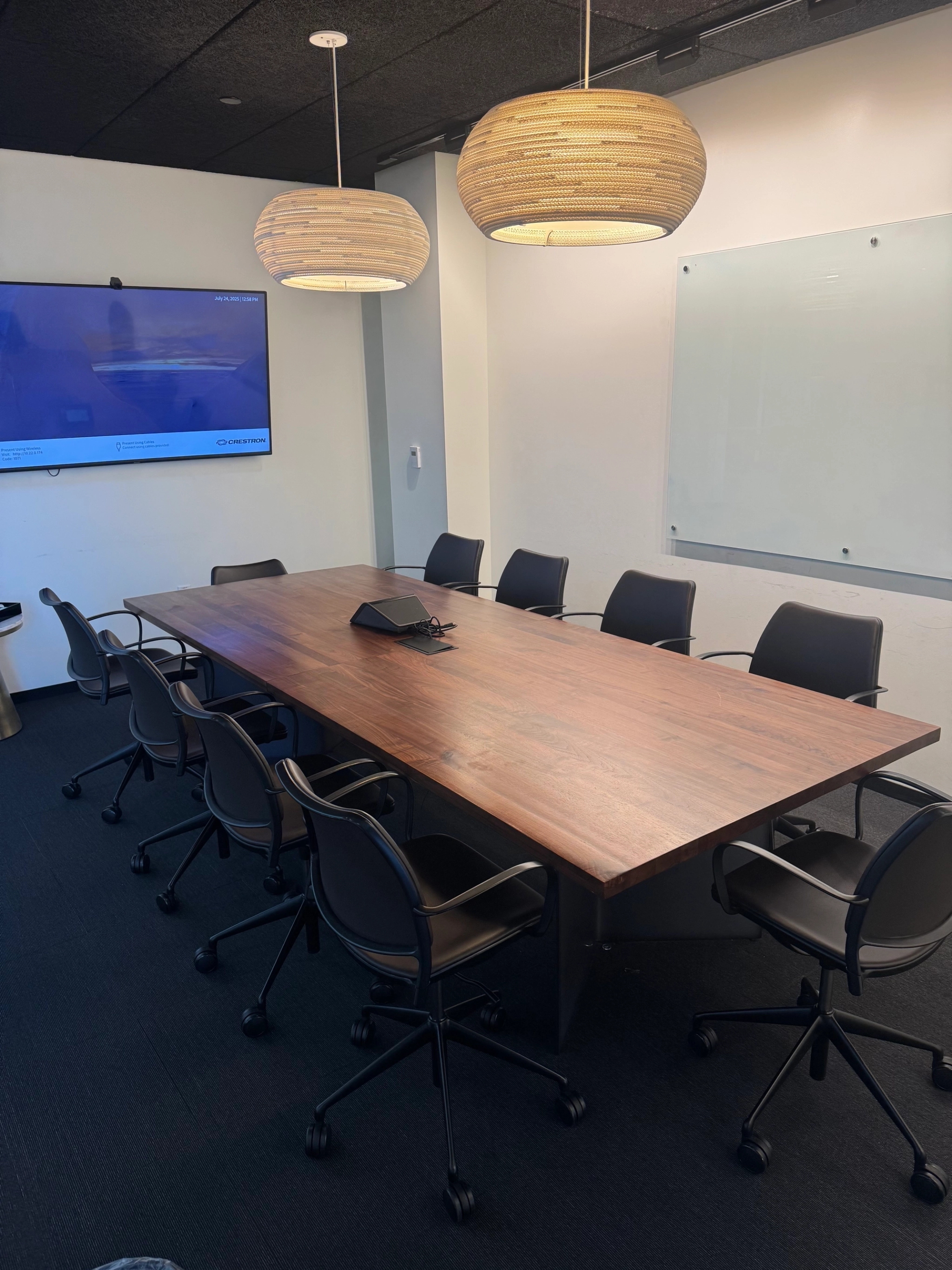 A modern coworking conference room with a wooden table, eight black chairs, a wall-mounted screen, two pendant lights, and a large whiteboard.