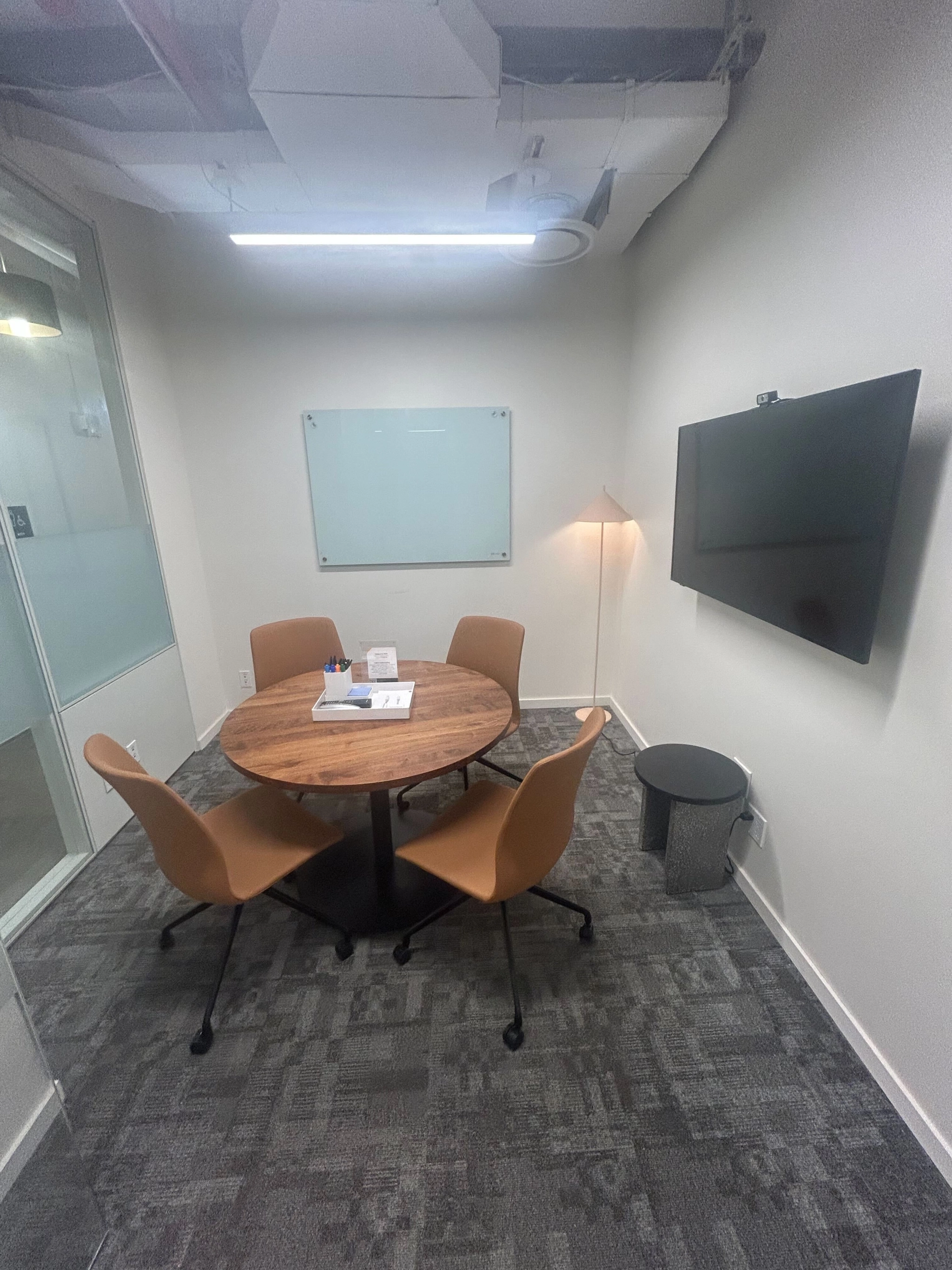 Small conference room in a New York coworking office, featuring a round table, four tan chairs, wall-mounted TV, glass whiteboard, and floor lamp. The space has gray carpet and white walls for a modern feel.