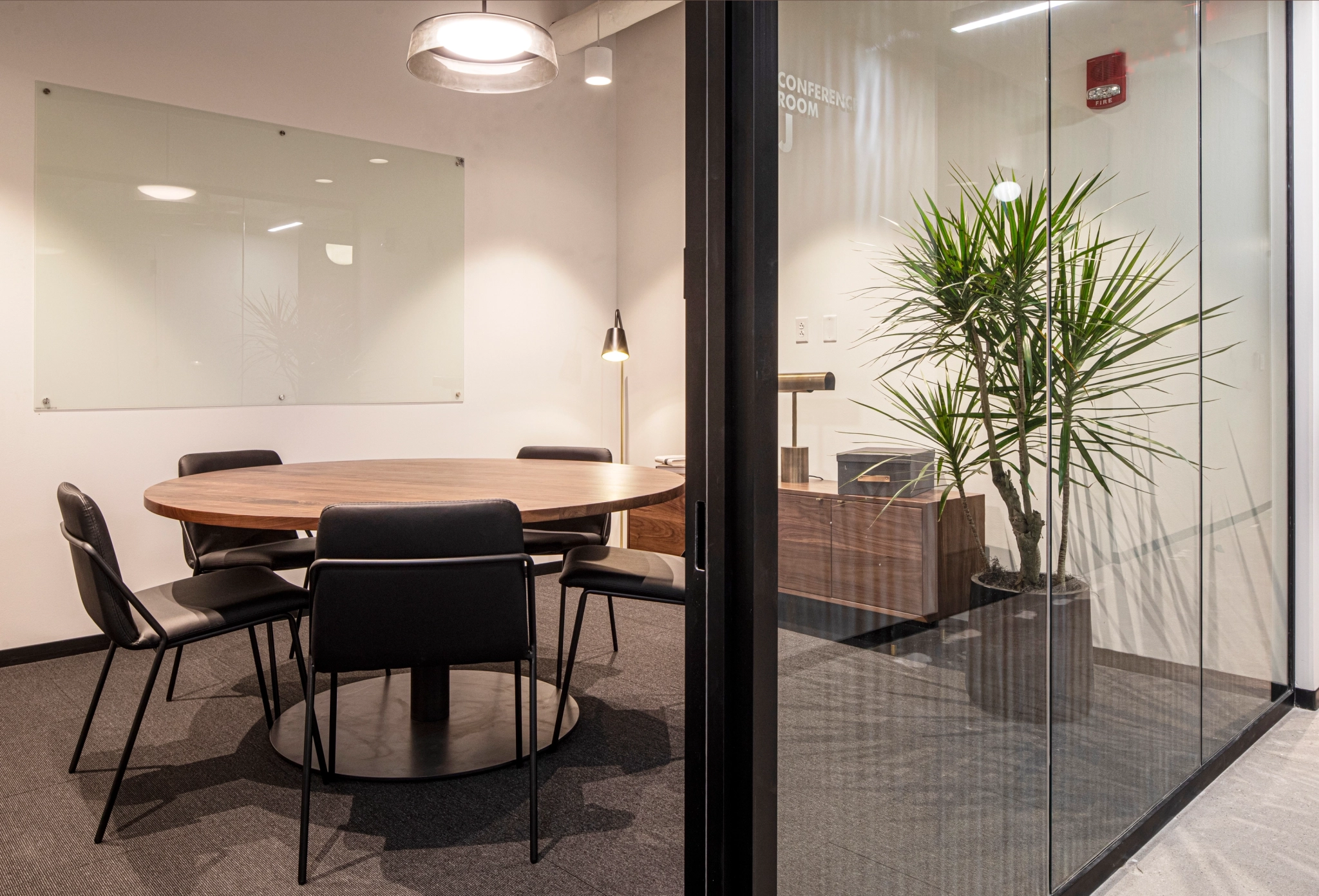A modern coworking meeting room with a round wooden table, six black chairs, a large glass board on the wall, and a tall plant near the glass wall.