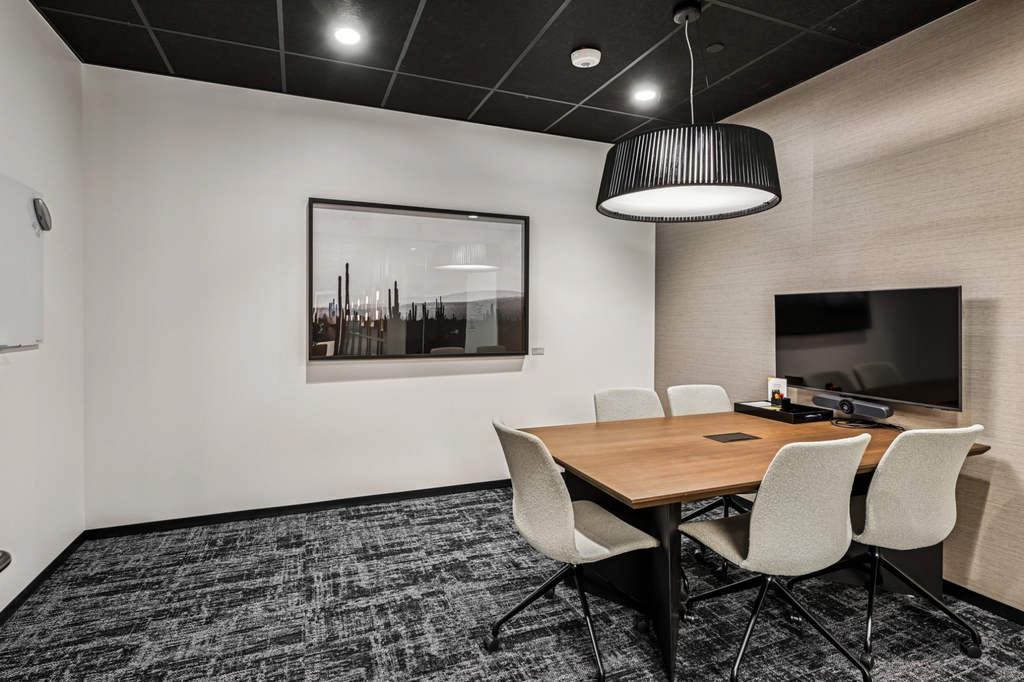 Modern coworking meeting room with a wooden table, four white chairs, wall-mounted TV, framed artwork, and a large overhead light fixture.