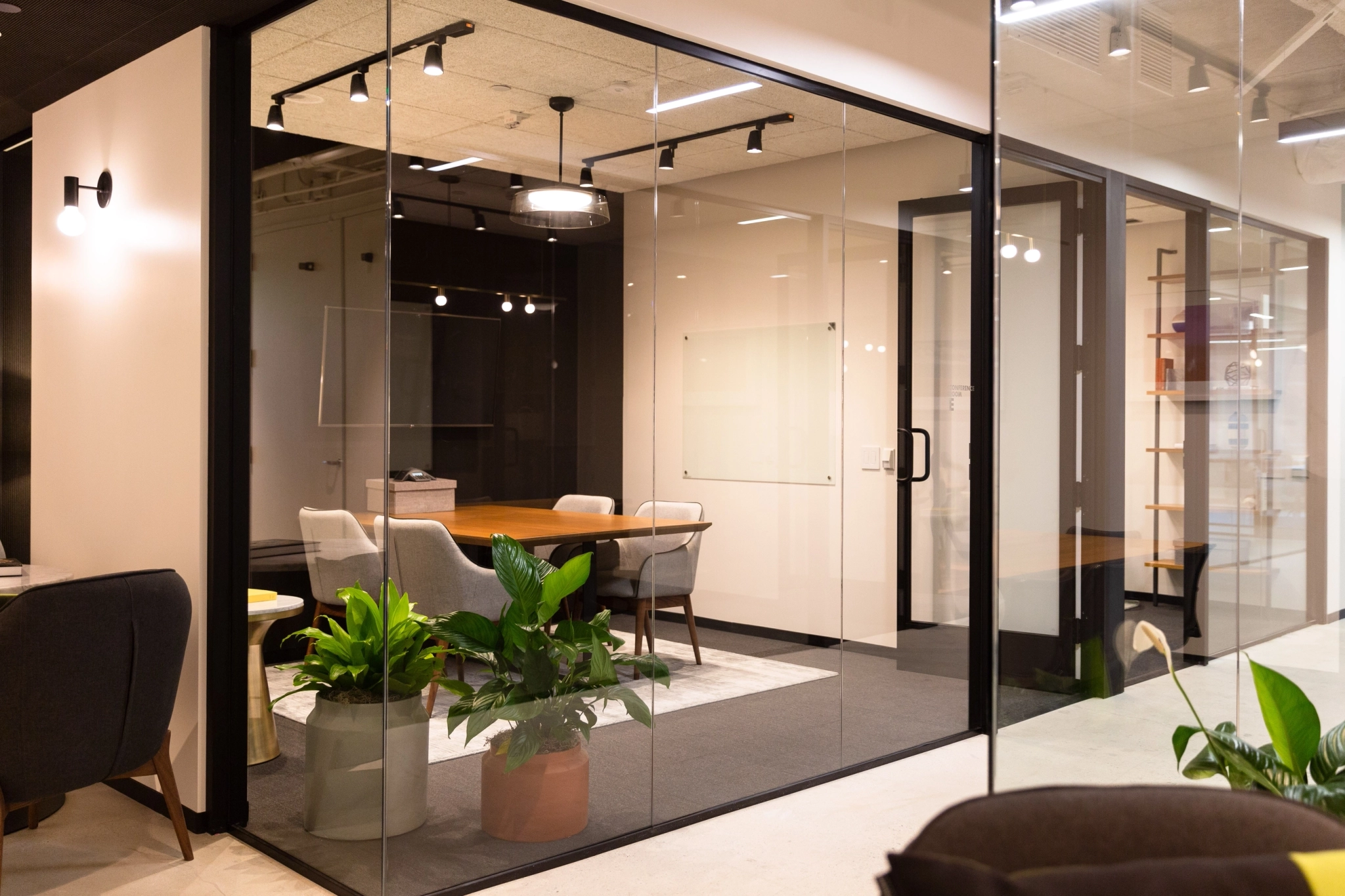 Modern coworking office meeting room with glass walls, a large table, several chairs, potted plants, and minimal decor.