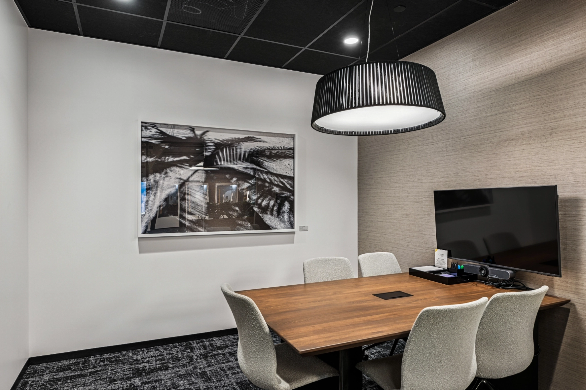 Modern meeting room in a coworking office, featuring a wooden table, six chairs, a wall-mounted TV, a large ceiling light, and abstract artwork on the wall.
