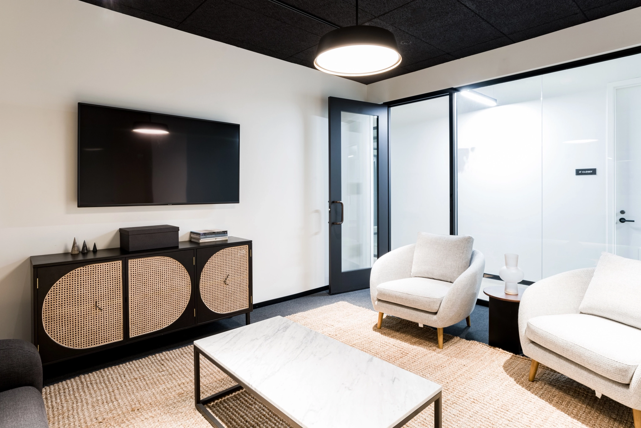 A modern office lounge with two armchairs, a sofa, a marble coffee table, a rug, and a TV mounted above a cabinet. A glass-walled meeting room is visible in the background, adding to the open workspace feel.