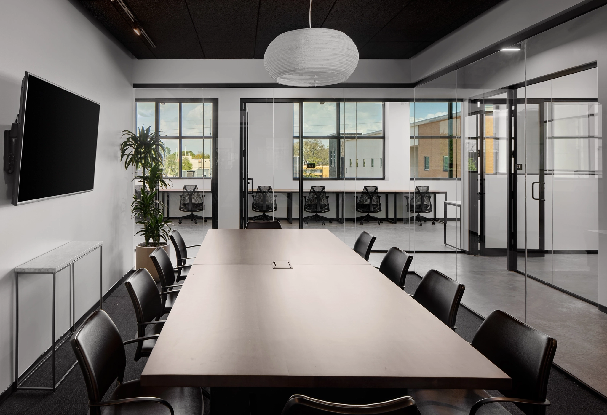 Modern Beverly Hills meeting room with a long wooden table, black chairs, wall-mounted TV, large pendant light, glass partitions, and windows overlooking an office building—a perfect workspace for collaboration.