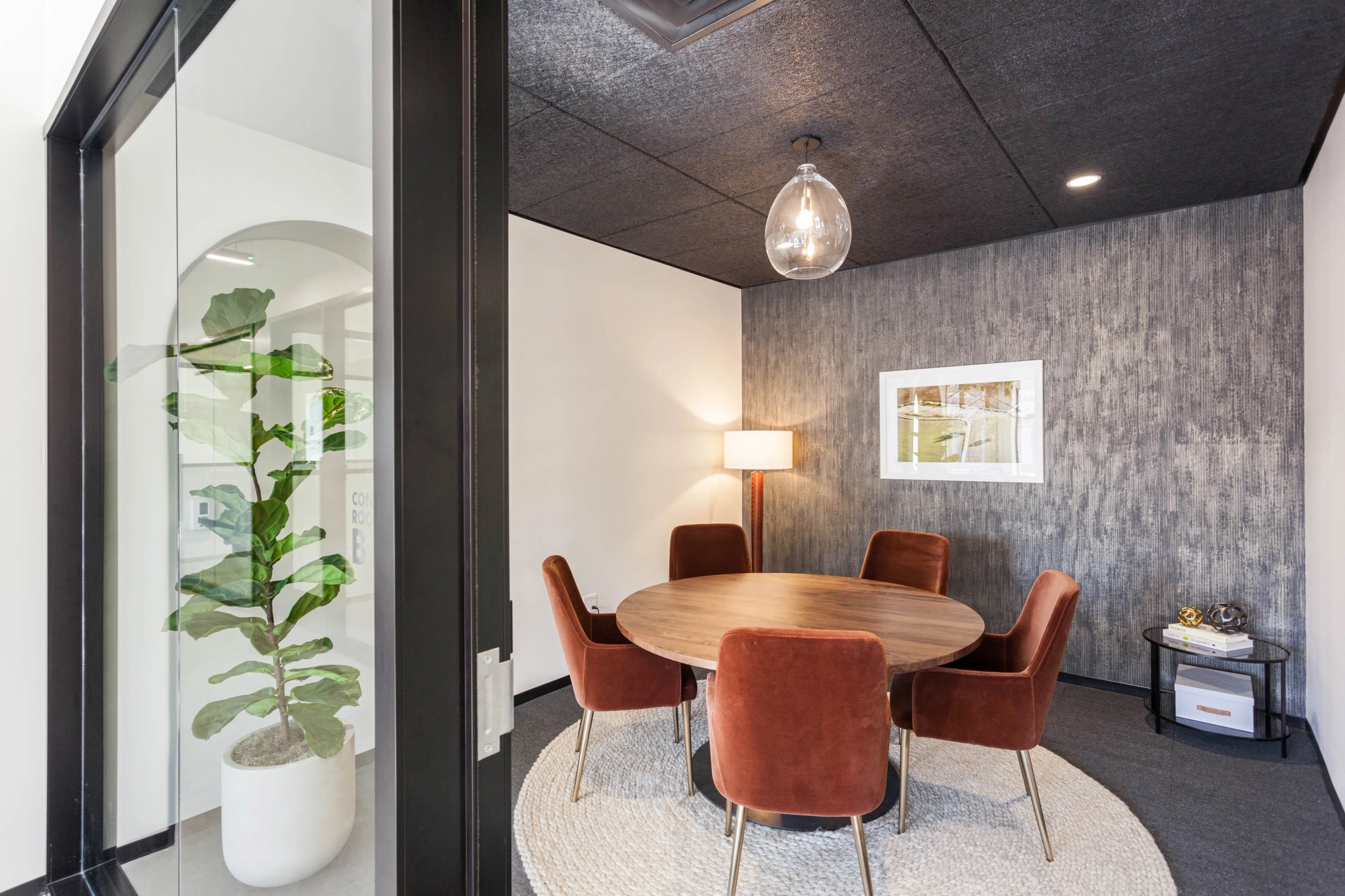 Cozy Beverly Hills meeting room with a round wooden table, four rust-colored chairs, a floor lamp, framed wall art, and a potted plant by the glass door—perfect for your next creative workspace session.