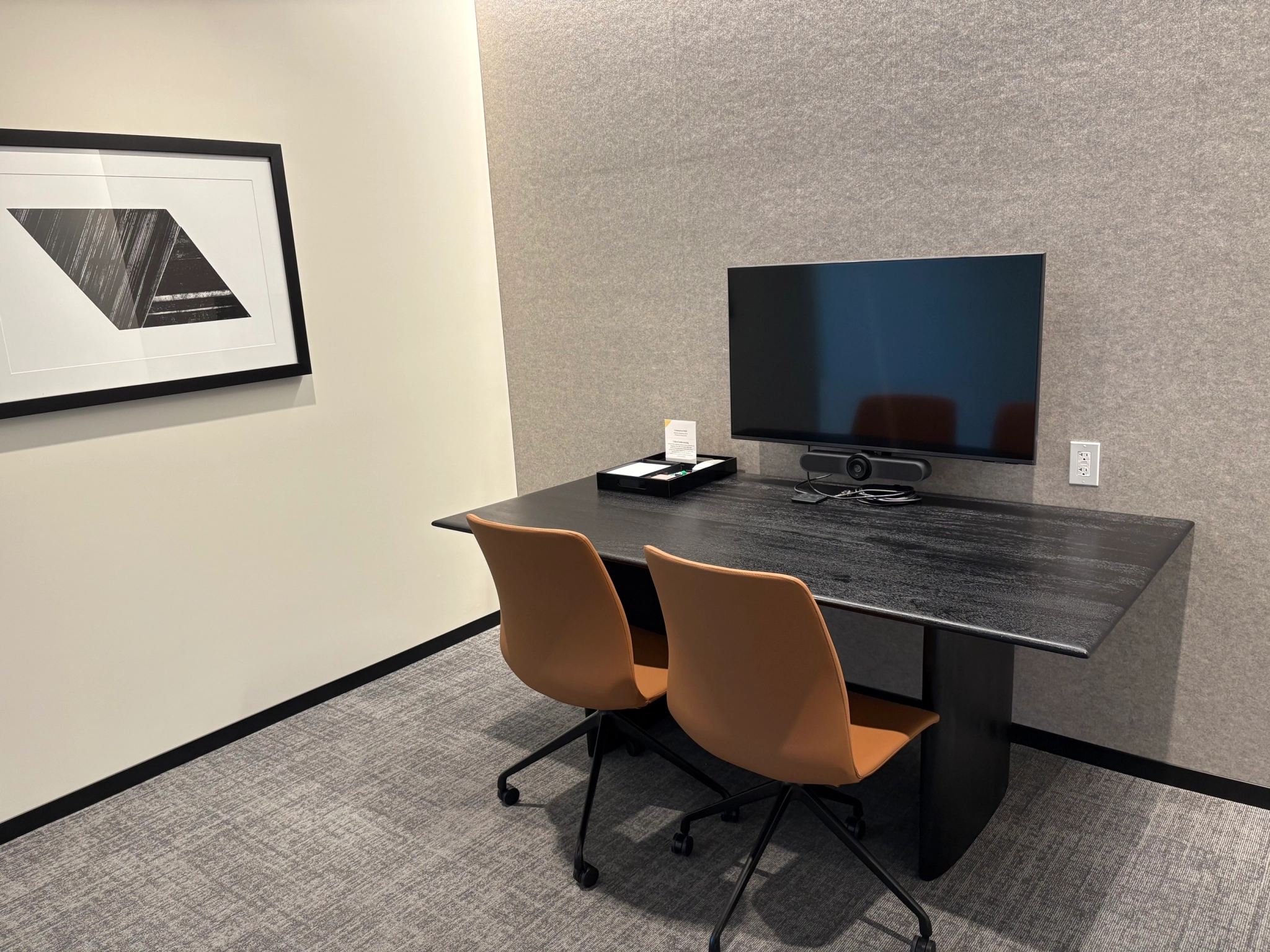 Cozy office meeting room with two brown chairs at a black table facing a monitor, framed abstract artwork on the wall, and gray carpet—ideal for focused work or small team discussions in your coworking space.
