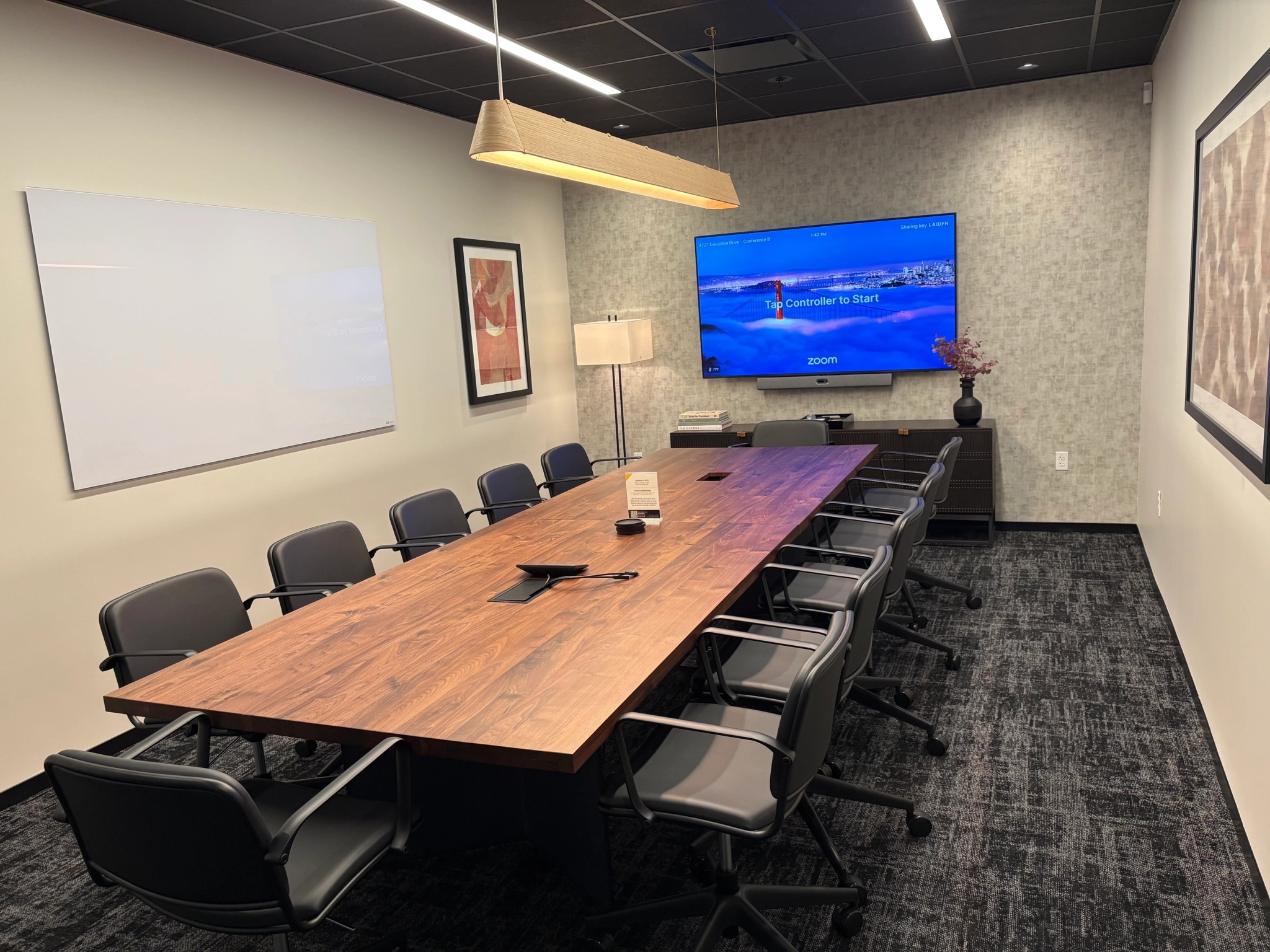 Modern coworking conference room in San Diego with a long wooden table, ten chairs, a wall-mounted screen displaying a Zoom interface, a whiteboard, and framed art on the walls.