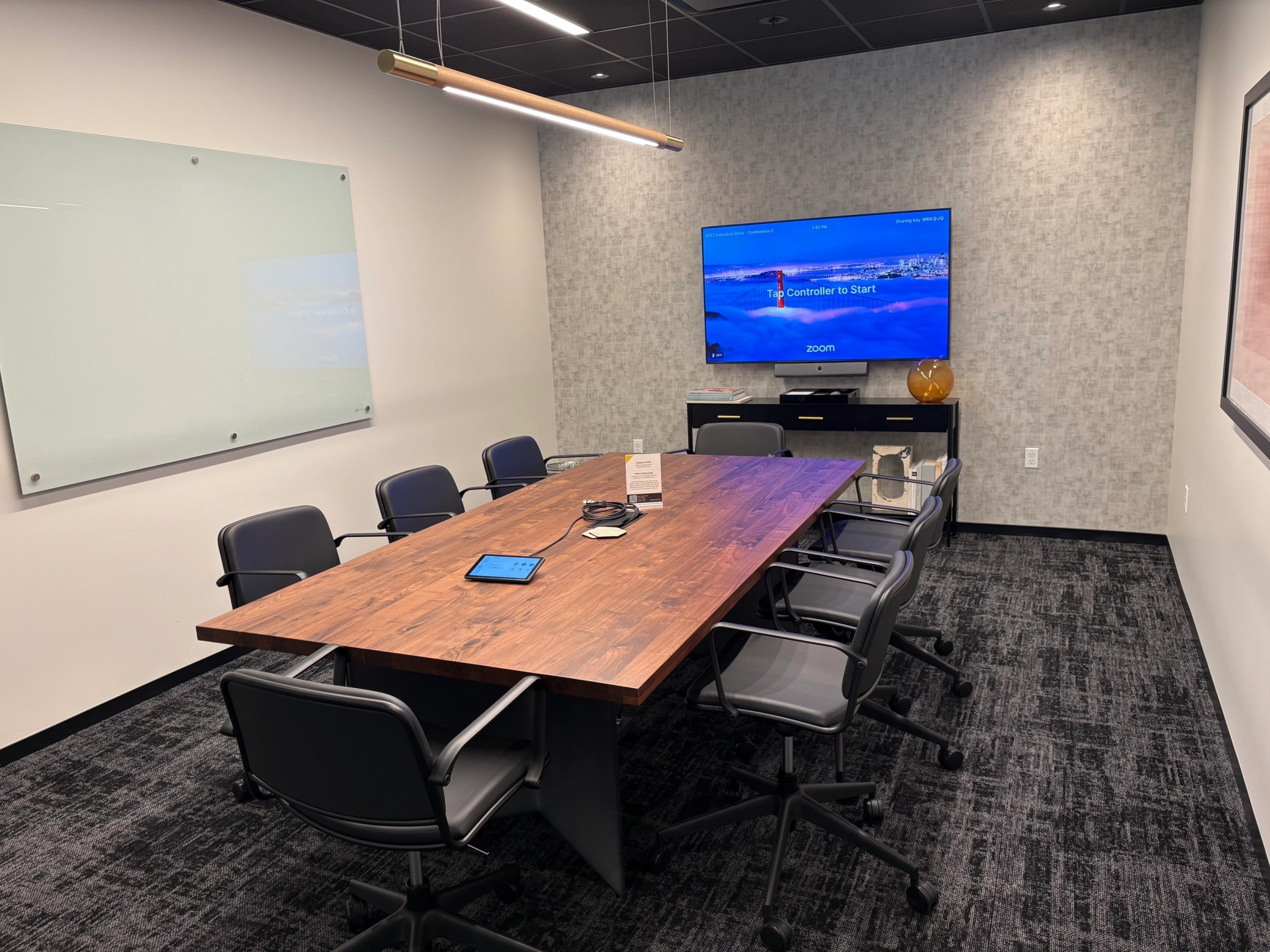 A modern office meeting room in San Diego with a wooden table, eight chairs, a wall-mounted screen displaying Zoom, a glass whiteboard, and a tablet on the table.