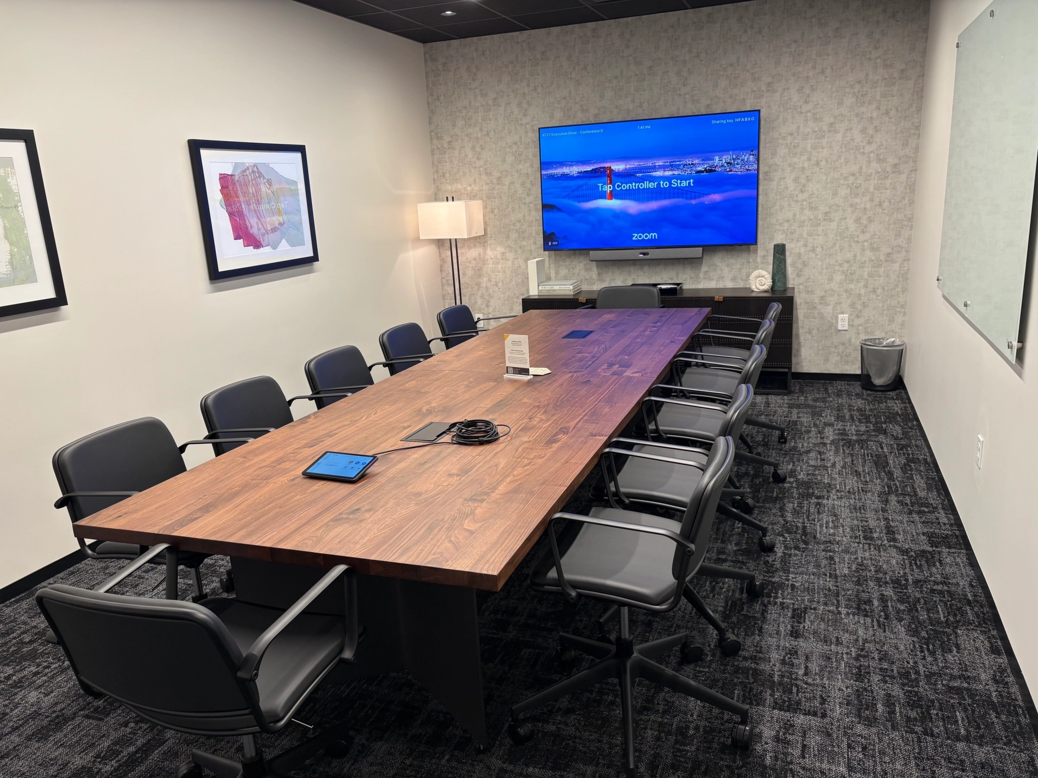 A modern coworking conference room in San Diego with a long wooden table, ten chairs, wall-mounted Zoom screen, framed art, a lamp, and a large glass whiteboard for an inspiring workspace.