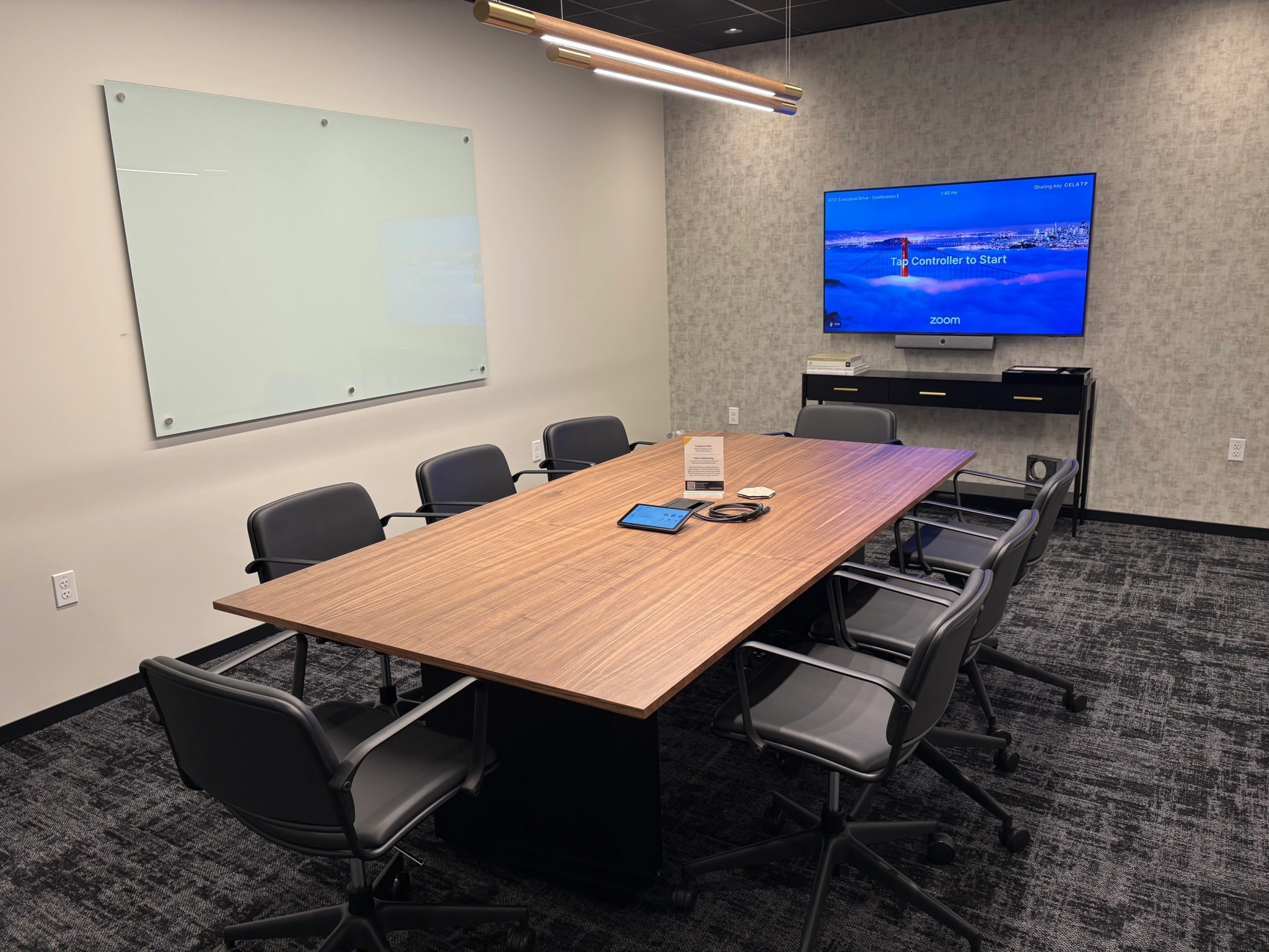 A modern San Diego workspace featuring a wooden table, eight chairs, a wall-mounted TV displaying a Zoom screen, and a glass whiteboard on the wall.