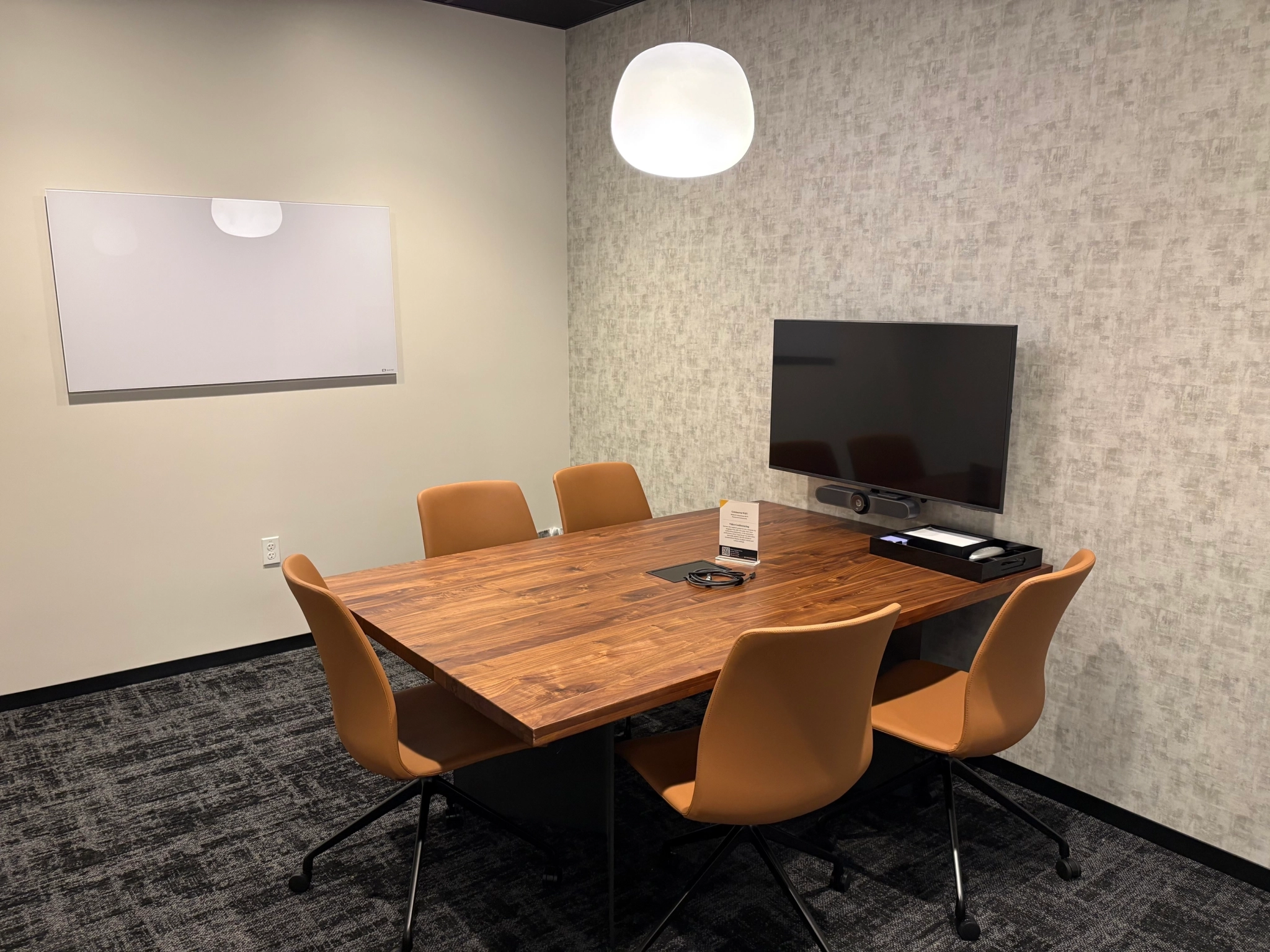 A small conference room in a San Diego coworking space features a wooden table, four tan chairs, a wall-mounted TV, video conferencing device, whiteboard, and a hanging light fixture—ideal for productive meetings.