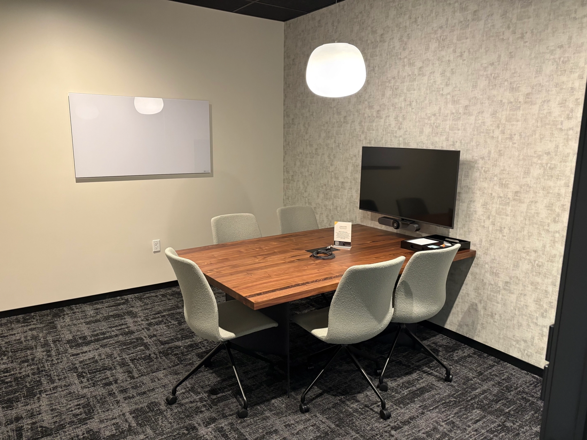 Coworking meeting room in San Diego with a wooden table, four gray chairs, a wall-mounted TV, a whiteboard, and a ceiling light.