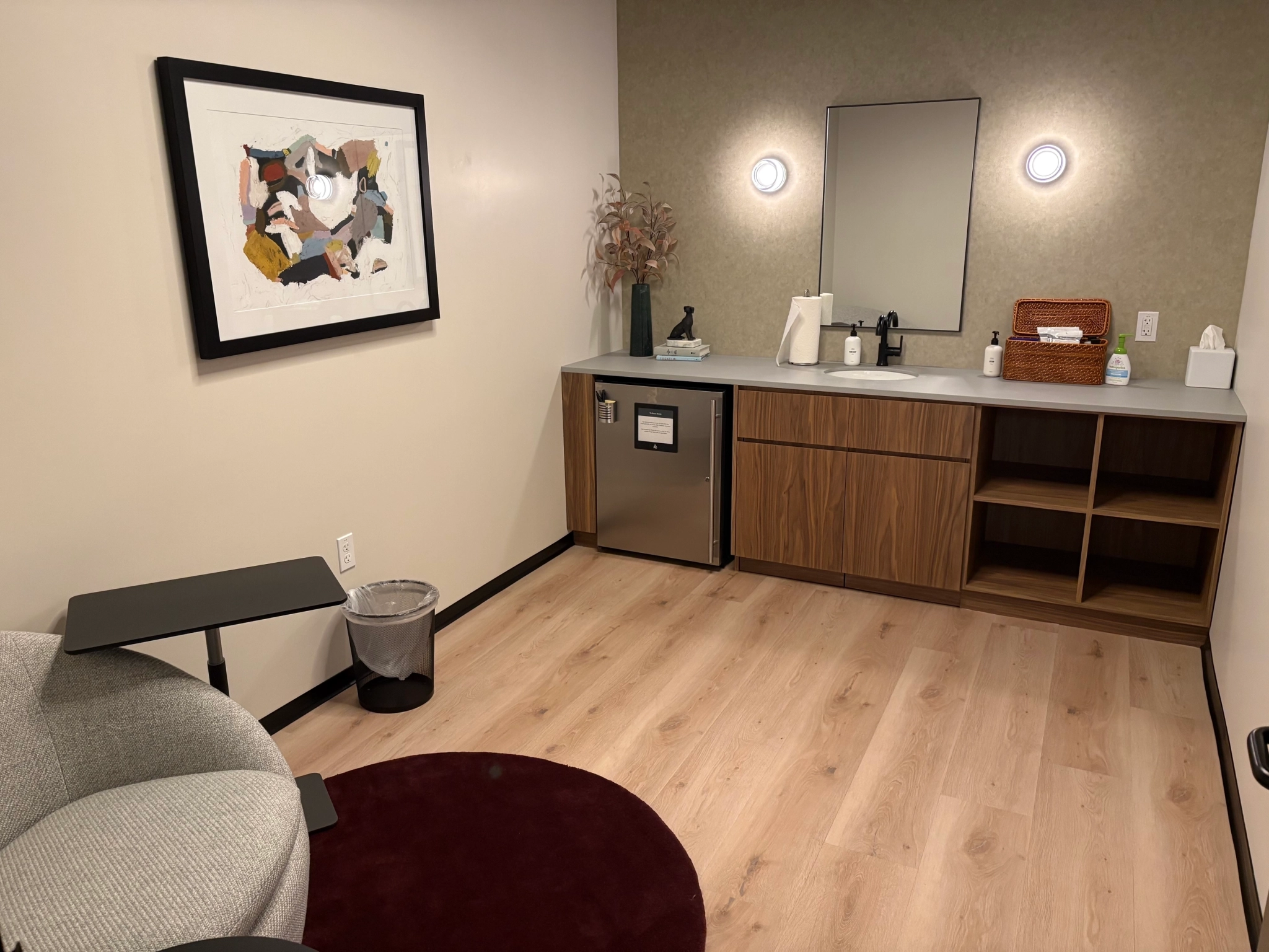 A small, modern coworking room in San Diego with a chair, side table, mini fridge, wooden cabinets, sink, mirror, wall art, and soft lighting.