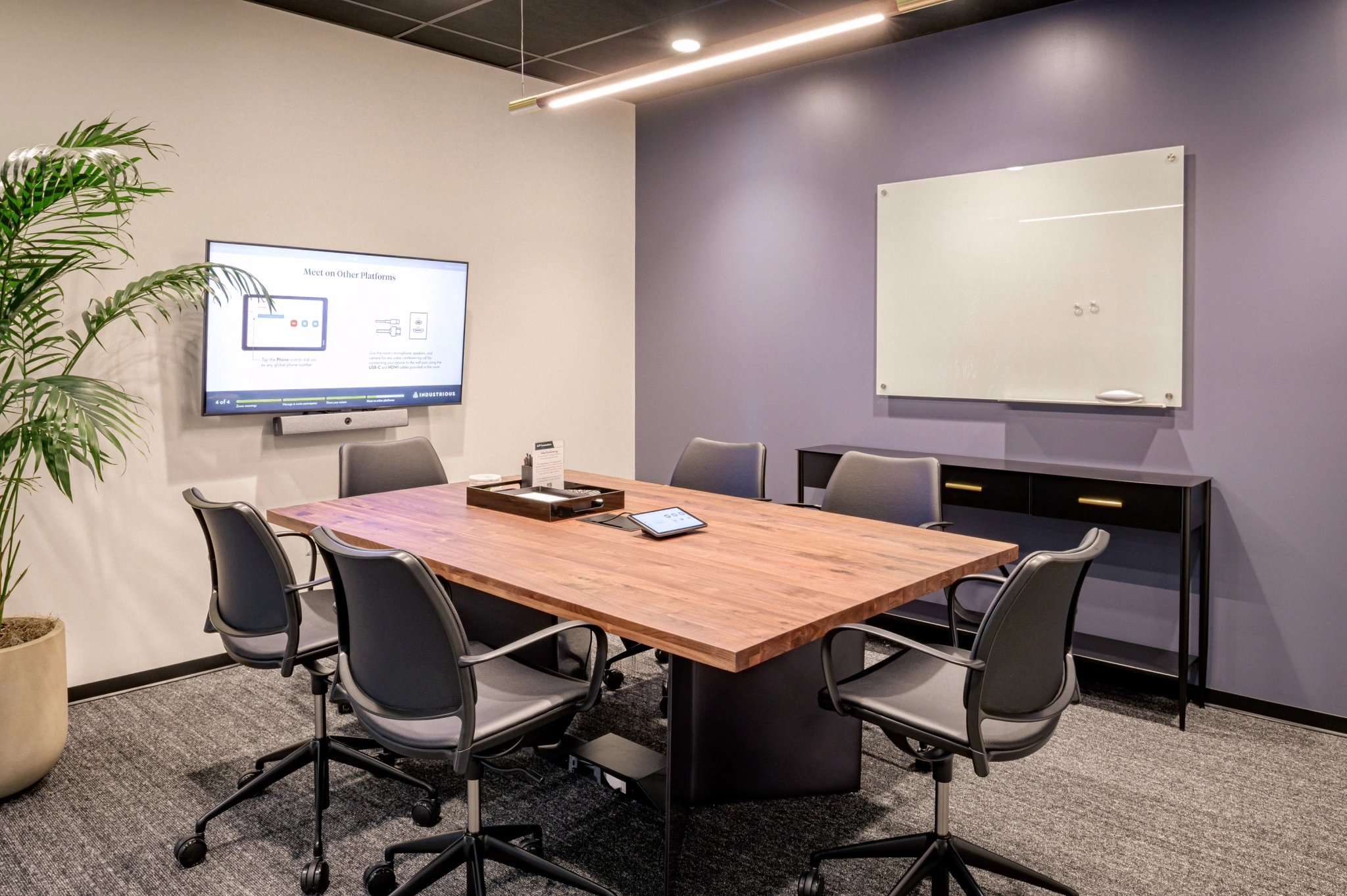 Modern meeting room in Toronto with a wooden table, five gray chairs, a wall-mounted screen displaying a presentation, a whiteboard, a credenza, and a potted plant in the corner—perfect for any workspace need.