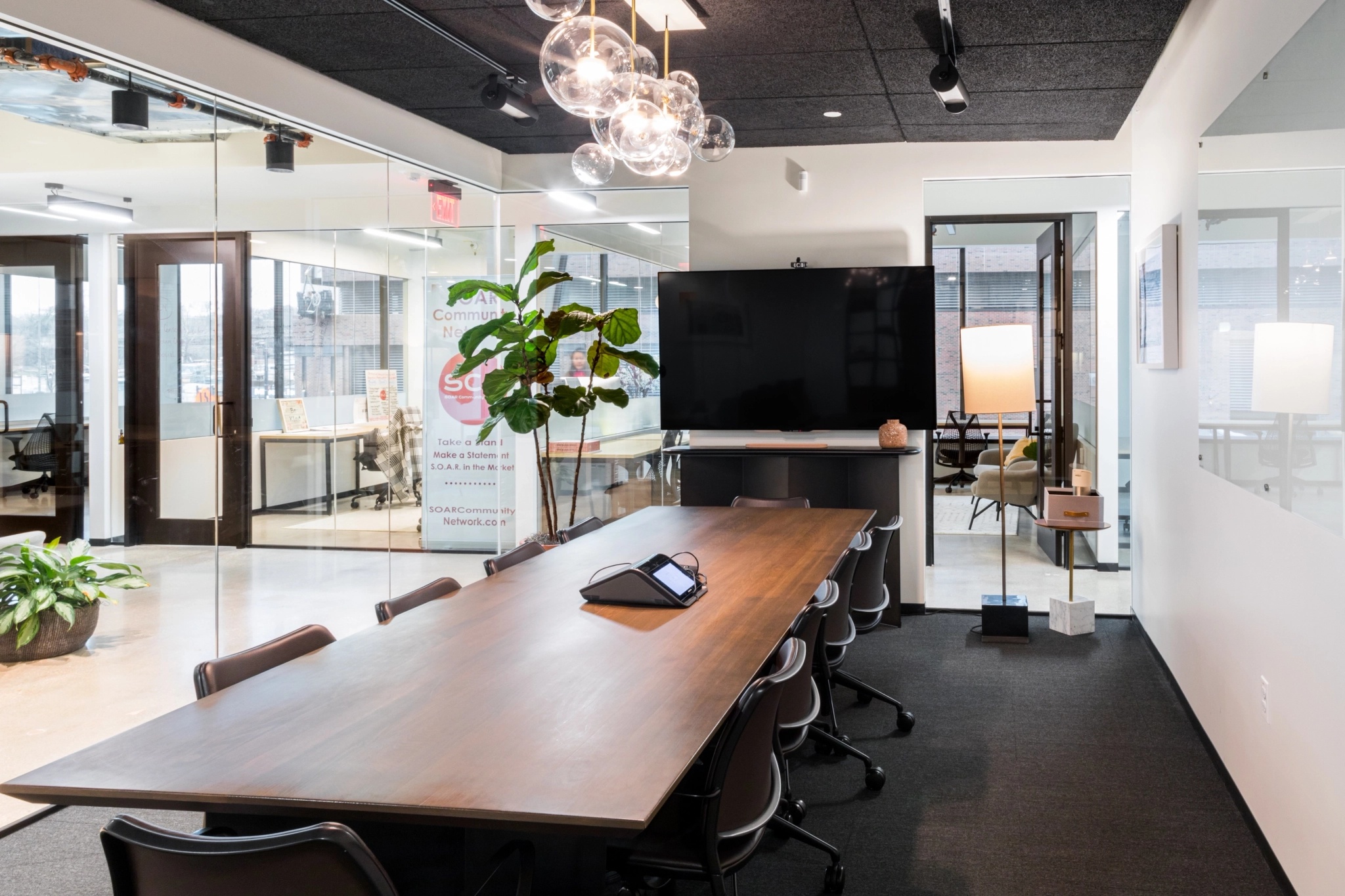 Modern meeting room with a long wooden table, several chairs, a large screen on the wall, glass walls, and pendant lights; an office area is visible through the glass in this stylish Austin coworking space.