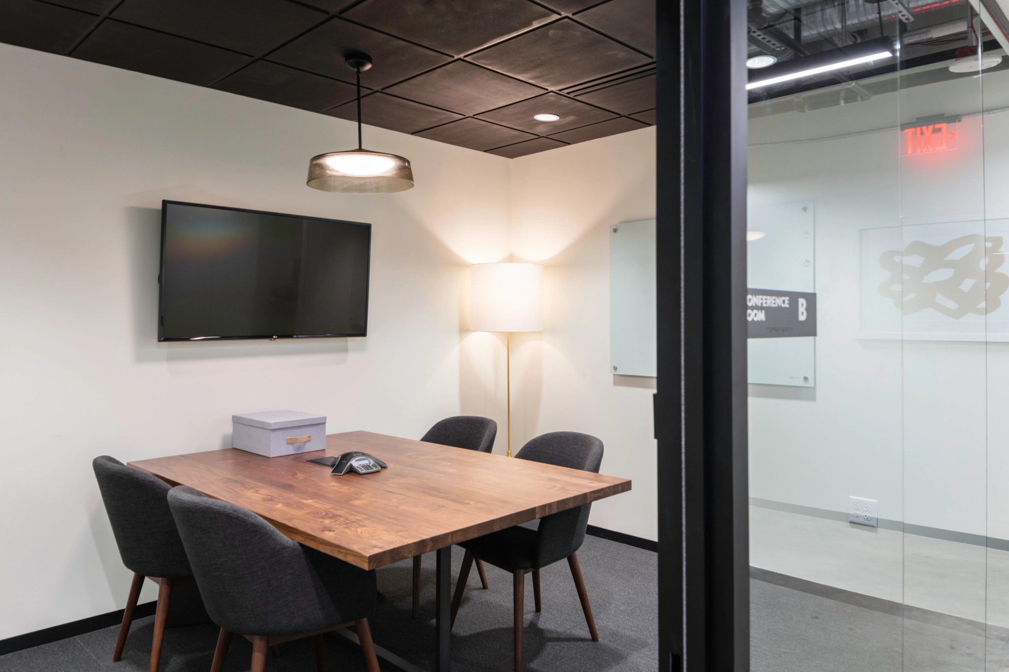 Small modern conference room in an Austin office, featuring a wooden table, four chairs, wall-mounted TV, lamp, and a box on the table; glass door partially open to workspace hallway.