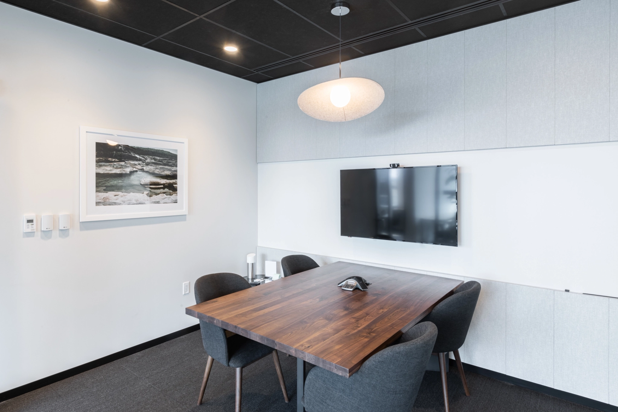 A small coworking conference room in Austin with a wooden table, four gray chairs, a wall-mounted TV, a conference phone, and a framed photo of waves on the wall.