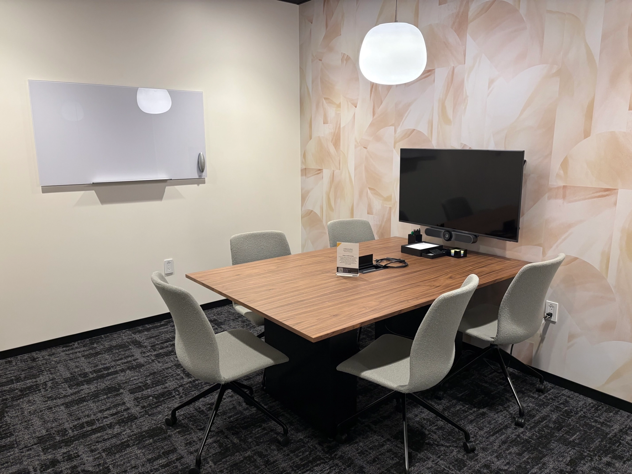 A small meeting room in East Palo Alto featuring a wooden table, six chairs, a wall-mounted TV, whiteboard, and soft lighting. The workspace is accented by walls with a subtle, abstract leaf pattern.