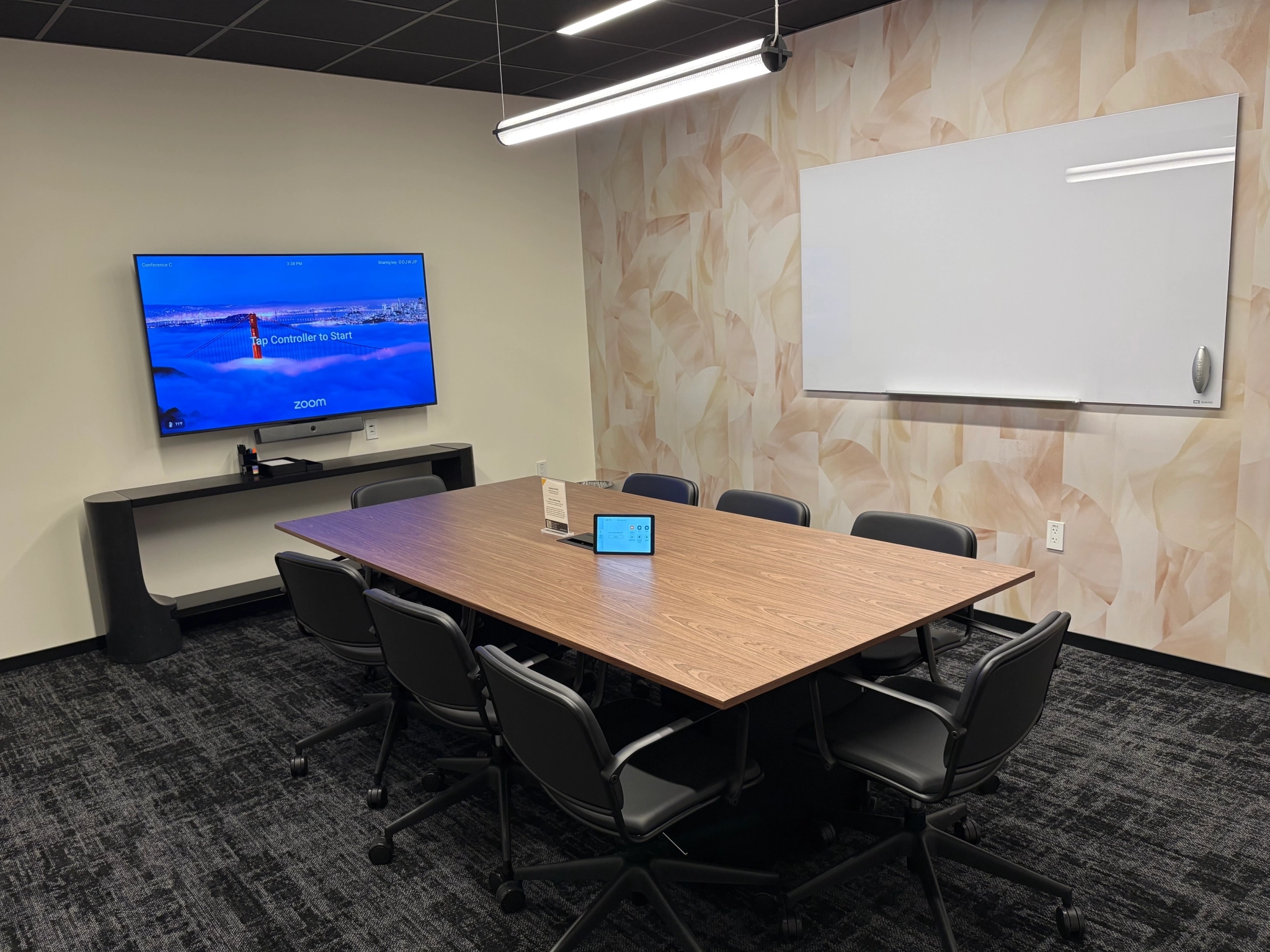 A coworking conference room with a wooden table, eight chairs, a wall-mounted TV displaying a Zoom screen, a whiteboard, and a touch panel on the table.