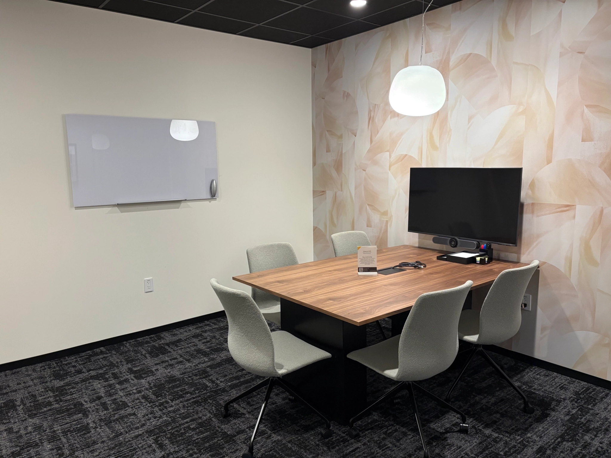 A small coworking meeting room with a table, four chairs, a wall-mounted screen, a whiteboard, and a pendant light, featuring patterned wallpaper on one wall.
