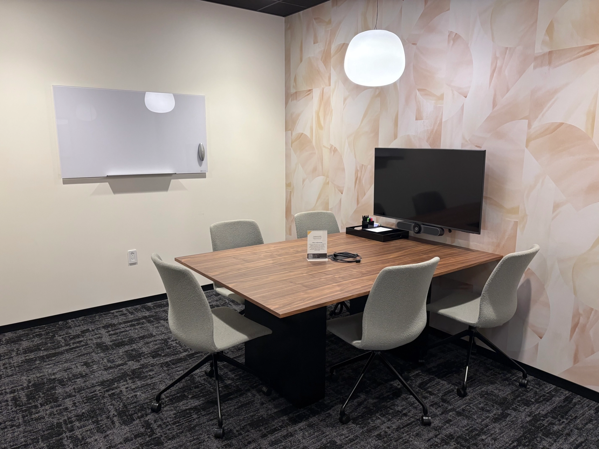 A small coworking meeting room with a wooden table, five gray chairs, a wall-mounted TV, a whiteboard, and a modern light fixture—perfect for your next workspace collaboration.