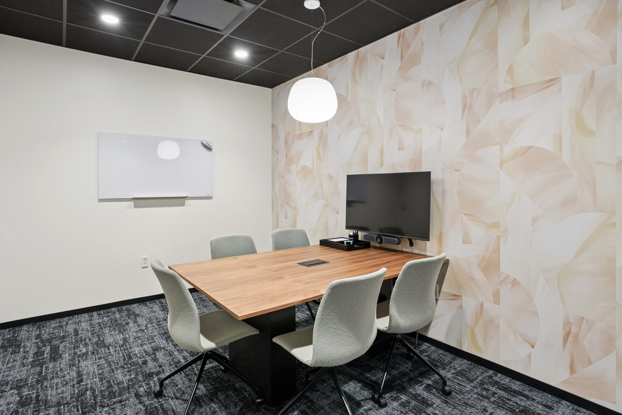 A small coworking meeting room with a table, five chairs, a wall-mounted screen, a whiteboard, and a pendant light, featuring patterned wallpaper on one wall.