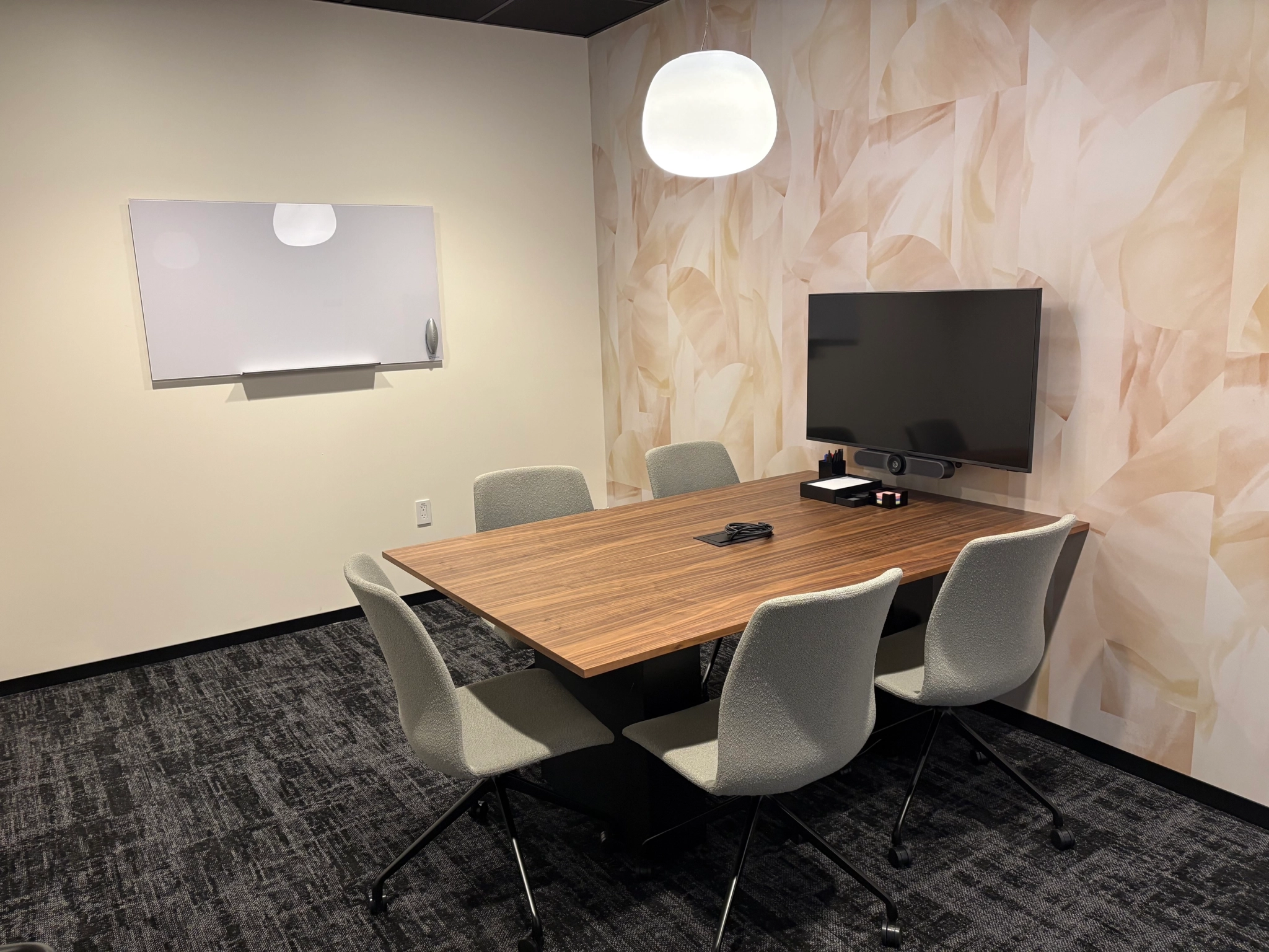 A small meeting room with a wooden table, five gray chairs, a wall-mounted TV, video conferencing equipment, a whiteboard, and a modern overhead light creates the perfect workspace for your team.