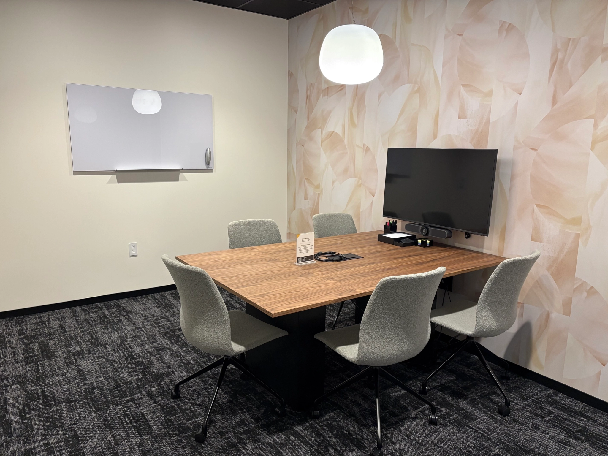 Une petite salle de réunion de bureau avec une table en bois, cinq chaises grises, un écran mural, un tableau blanc et une lampe suspendue.