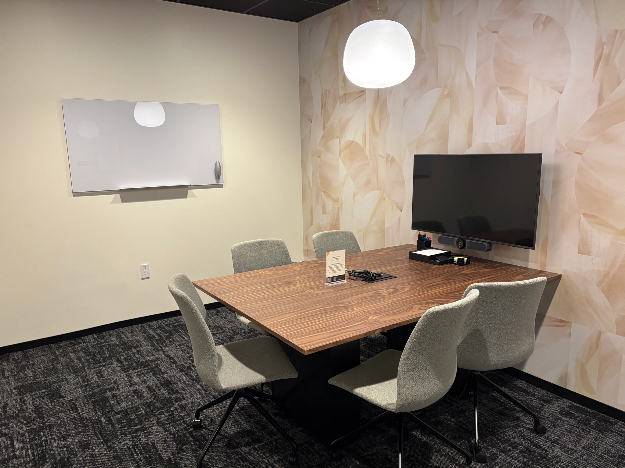 A small meeting room in East Palo Alto featuring a wooden table, four gray chairs, a wall-mounted monitor, a whiteboard, and a pendant light—ideal for focused work sessions or collaborative meetings.