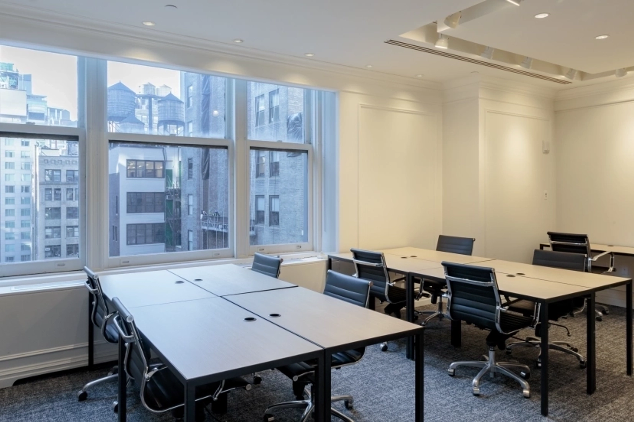 Modern workspace featuring several empty desks and black chairs, large windows overlooking New York city buildings, and neutral-colored walls and carpeting.