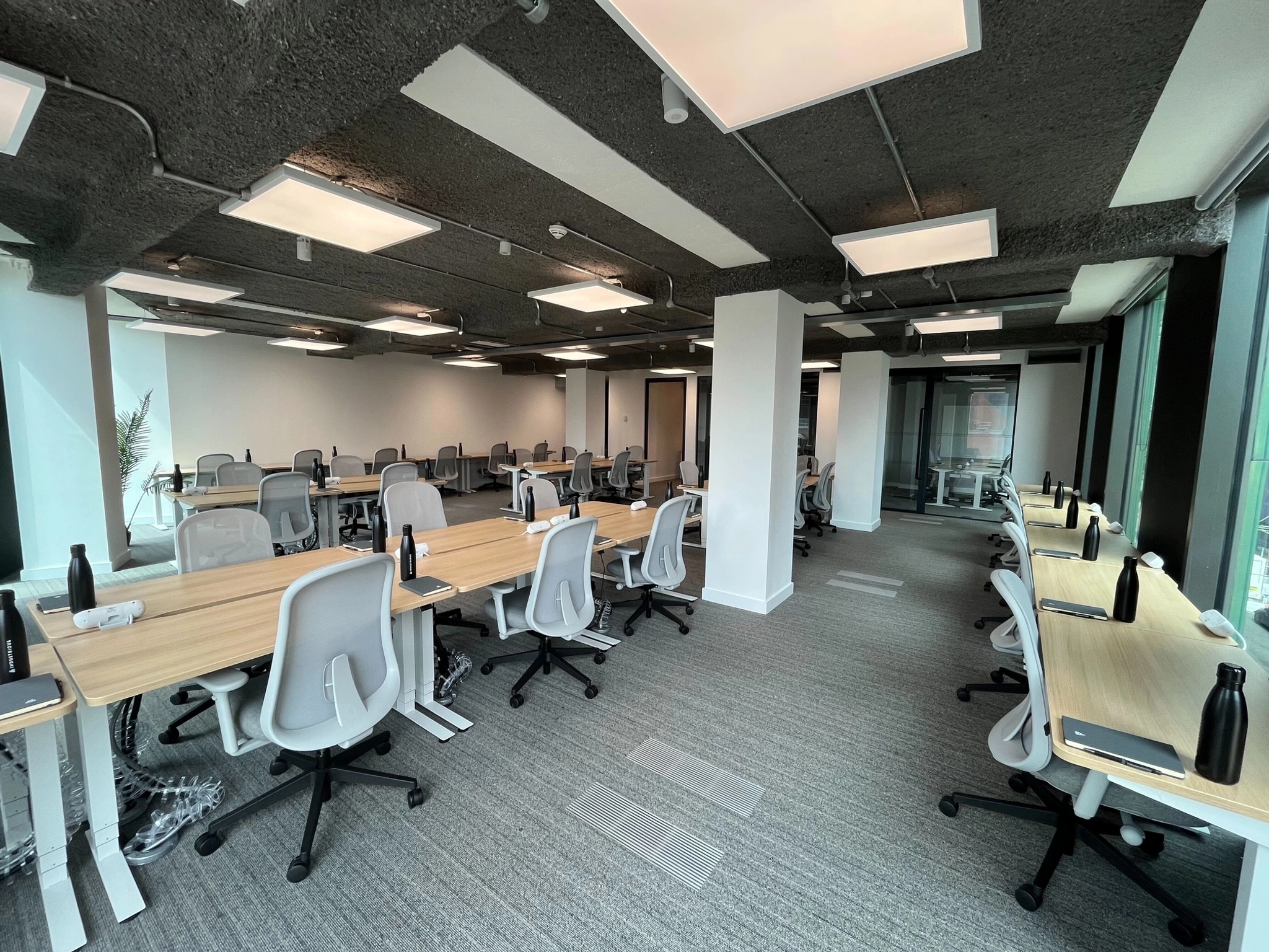 Modern open-plan workspace in Manchester with rows of empty desks and chairs, water bottles, and large windows providing natural light; ideal for collaboration or accessing a nearby meeting room.