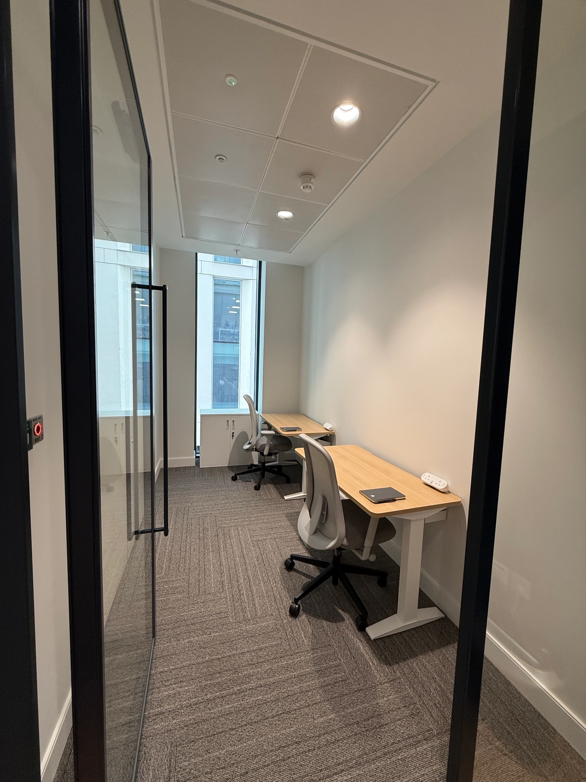 A small workspace with two desks and chairs, a window at the end, and neutral-colored walls and carpet—ideal for focused coworking or private meetings.