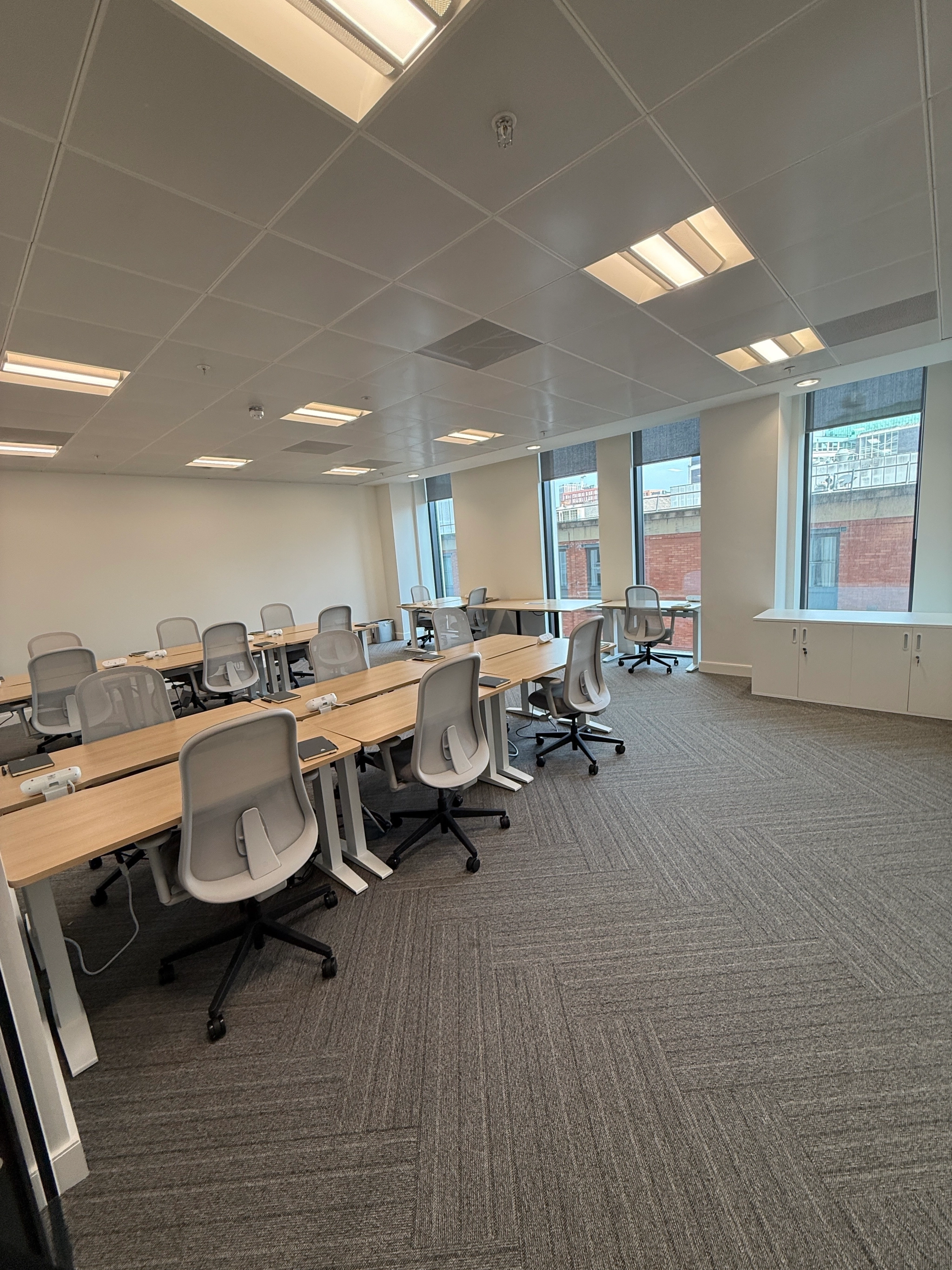 A modern coworking office space with empty desks and chairs, large windows, neutral colors, and overhead fluorescent lighting.