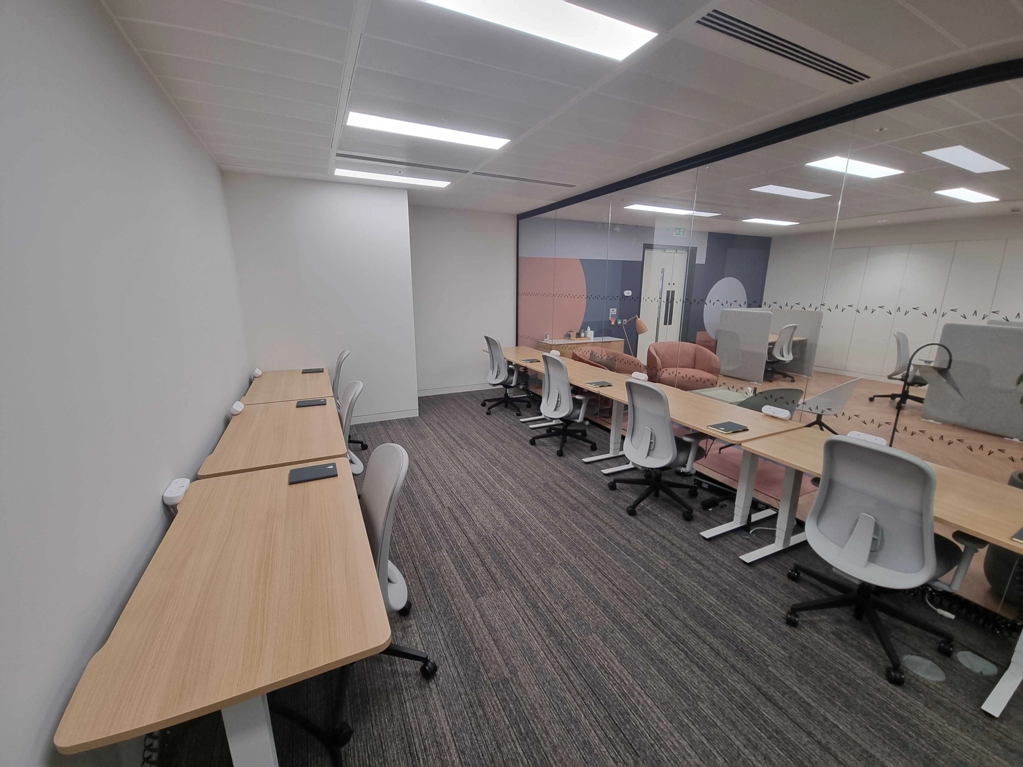 A modern office workspace with empty desks and chairs, white walls, carpeting, and a glass partition separating two meeting rooms.