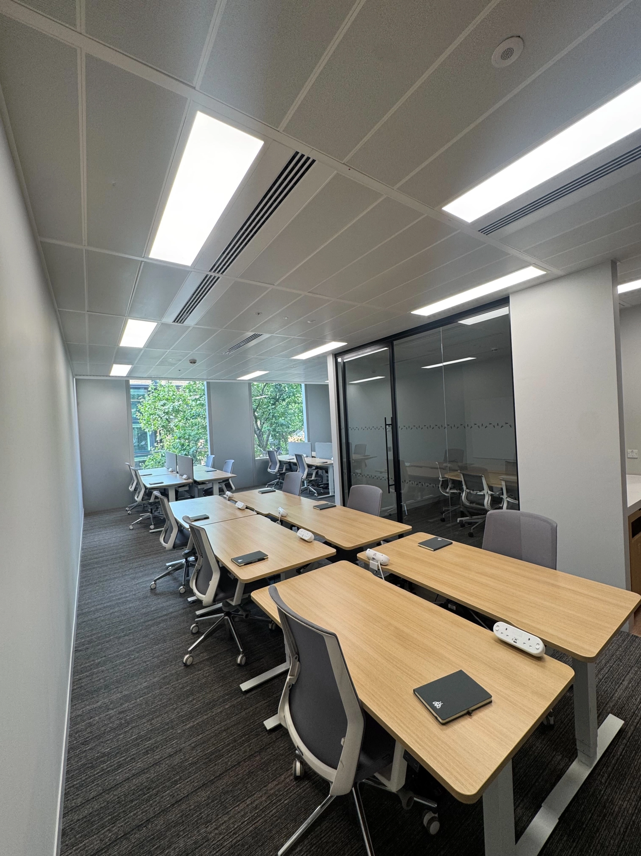Modern coworking office space with several wooden desks, gray chairs, notepads, and power strips, featuring large windows and a glass partition, with trees visible outside.