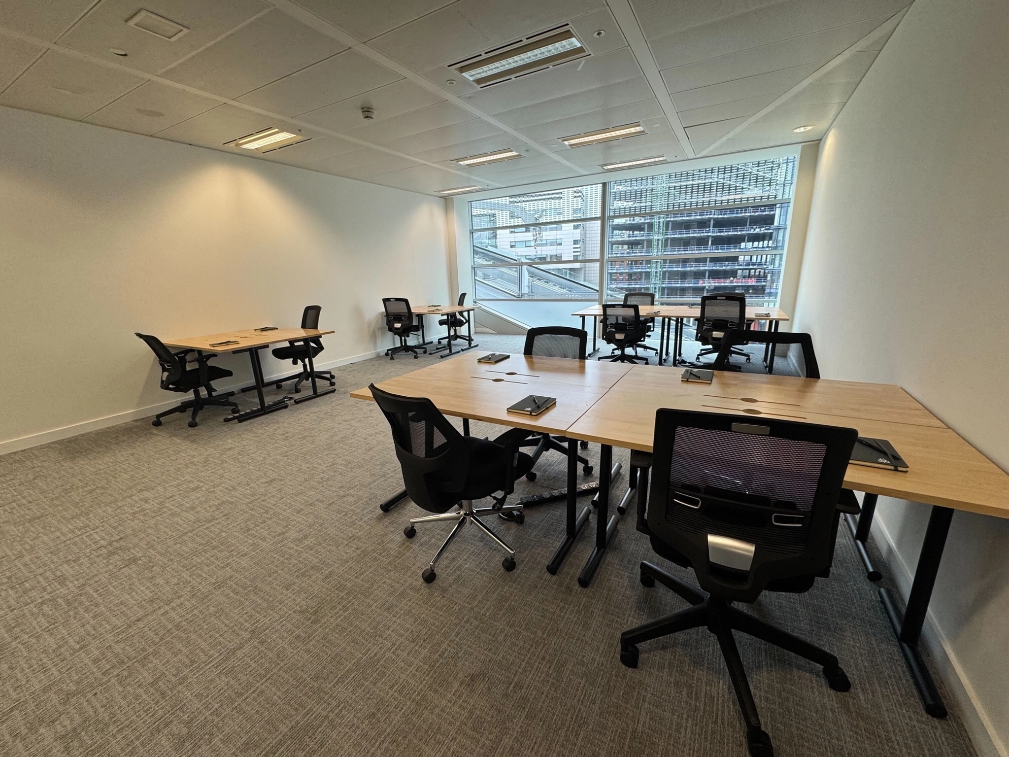 A modern coworking office room in London with six desks and chairs, notepads on each desk, gray carpet, white walls, and large windows overlooking a cityscape.