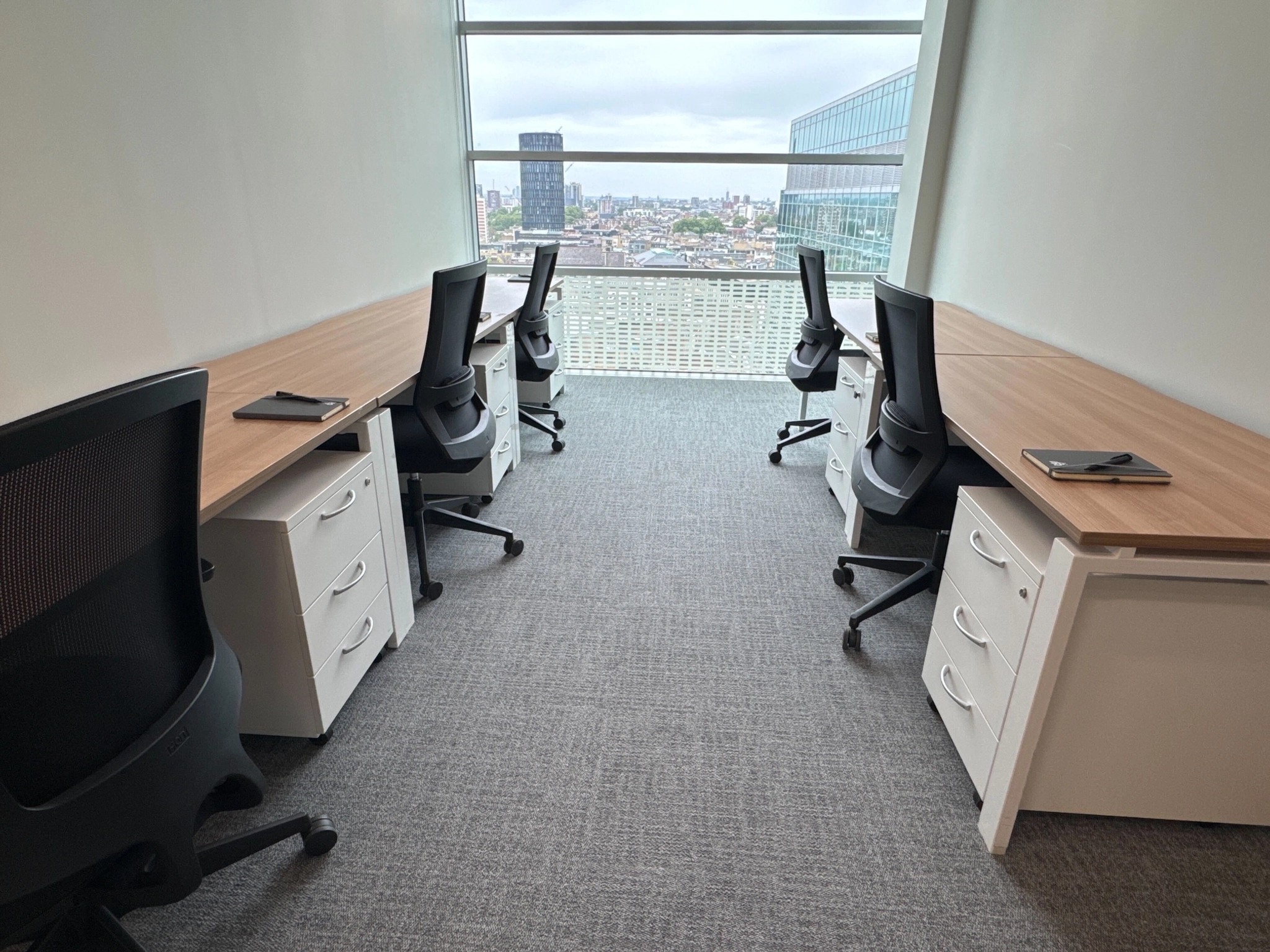 A modern London workspace featuring four desks, four black office chairs, and a large window overlooking a cityscape. Each desk has a closed notebook and a pen, making it perfect for use as a meeting room.
