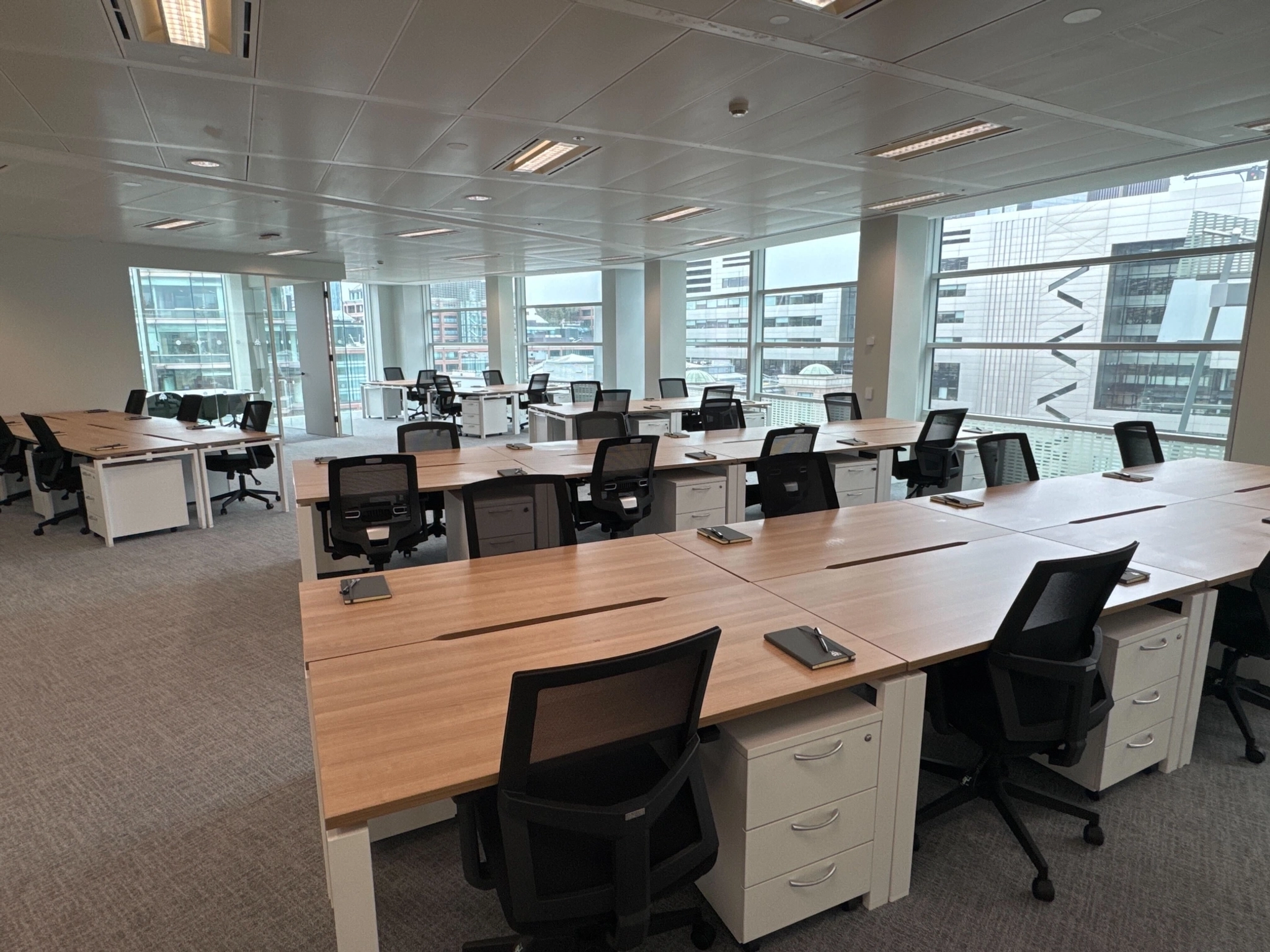 Espace de travail ouvert et moderne avec plusieurs bureaux et chaises vides, de grandes fenêtres et des bâtiments de la ville visibles de l'extérieur - parfait pour le coworking dans un cadre professionnel et lumineux.