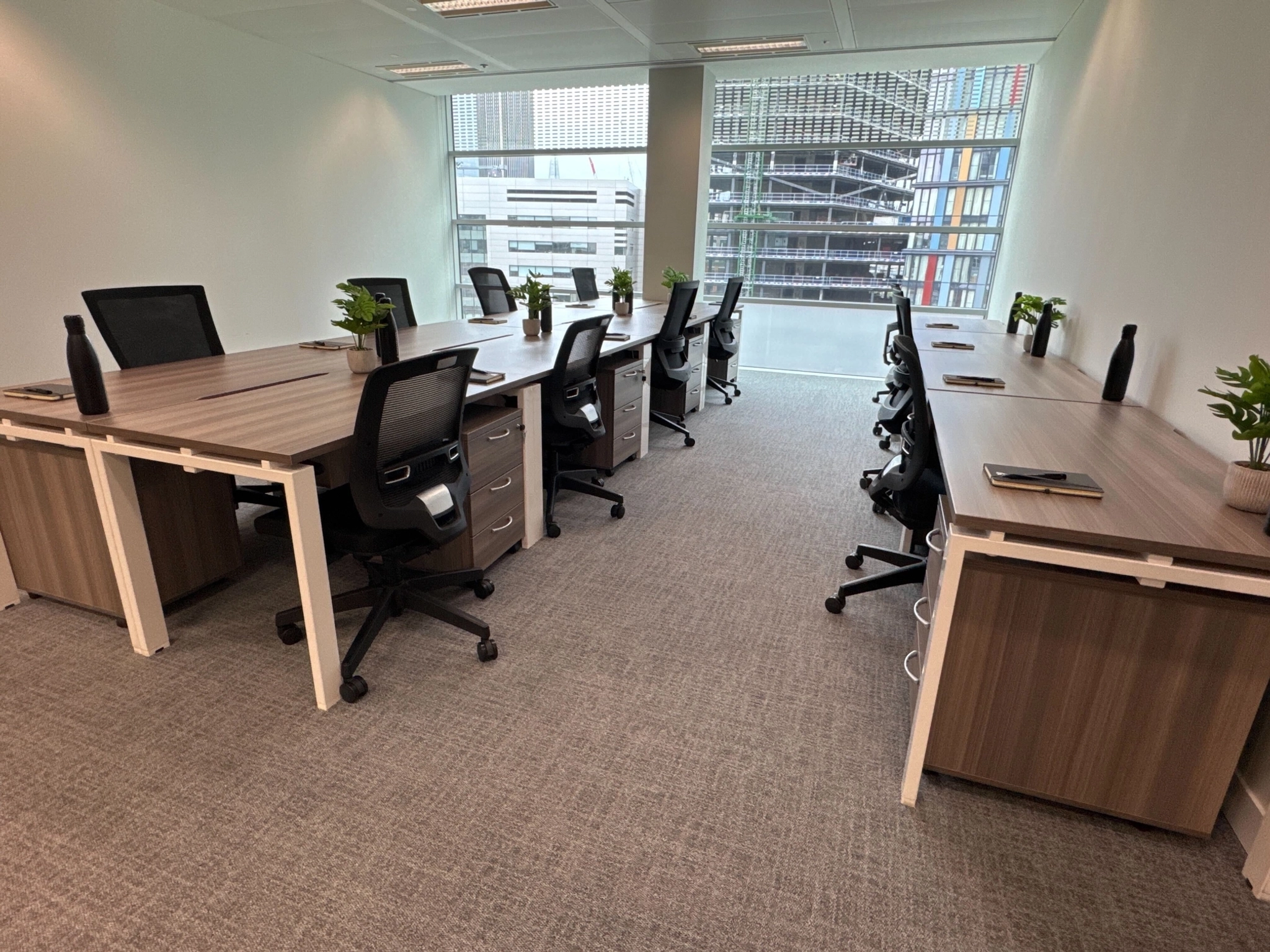 Modern coworking office with two rows of desks, ergonomic chairs, water bottles, notebooks, and potted plants, set near a large window overlooking London city buildings.