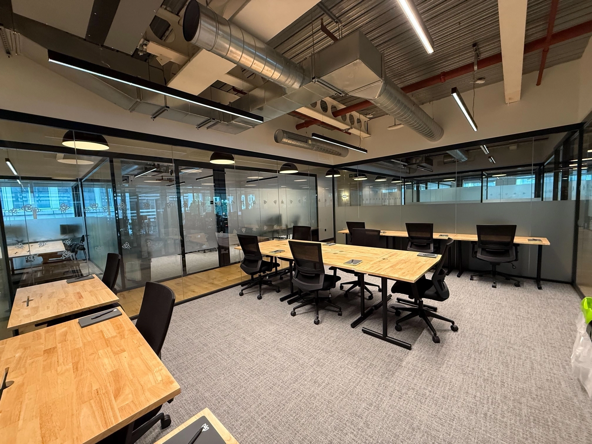 Modern open-plan office in London featuring wooden desks, black chairs, glass partitions, and exposed ceiling pipes. This well-lit workspace is mostly unoccupied, offering a bright and inviting environment.