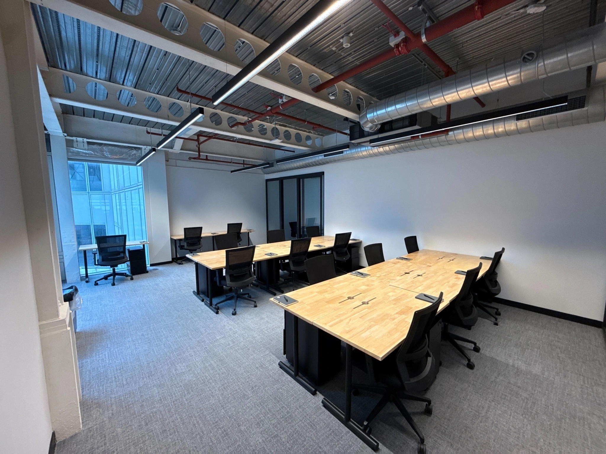 Modern London office space with multiple wooden desks, black office chairs, and large windows. The workspace features exposed ceilings with ducts and overhead lighting.