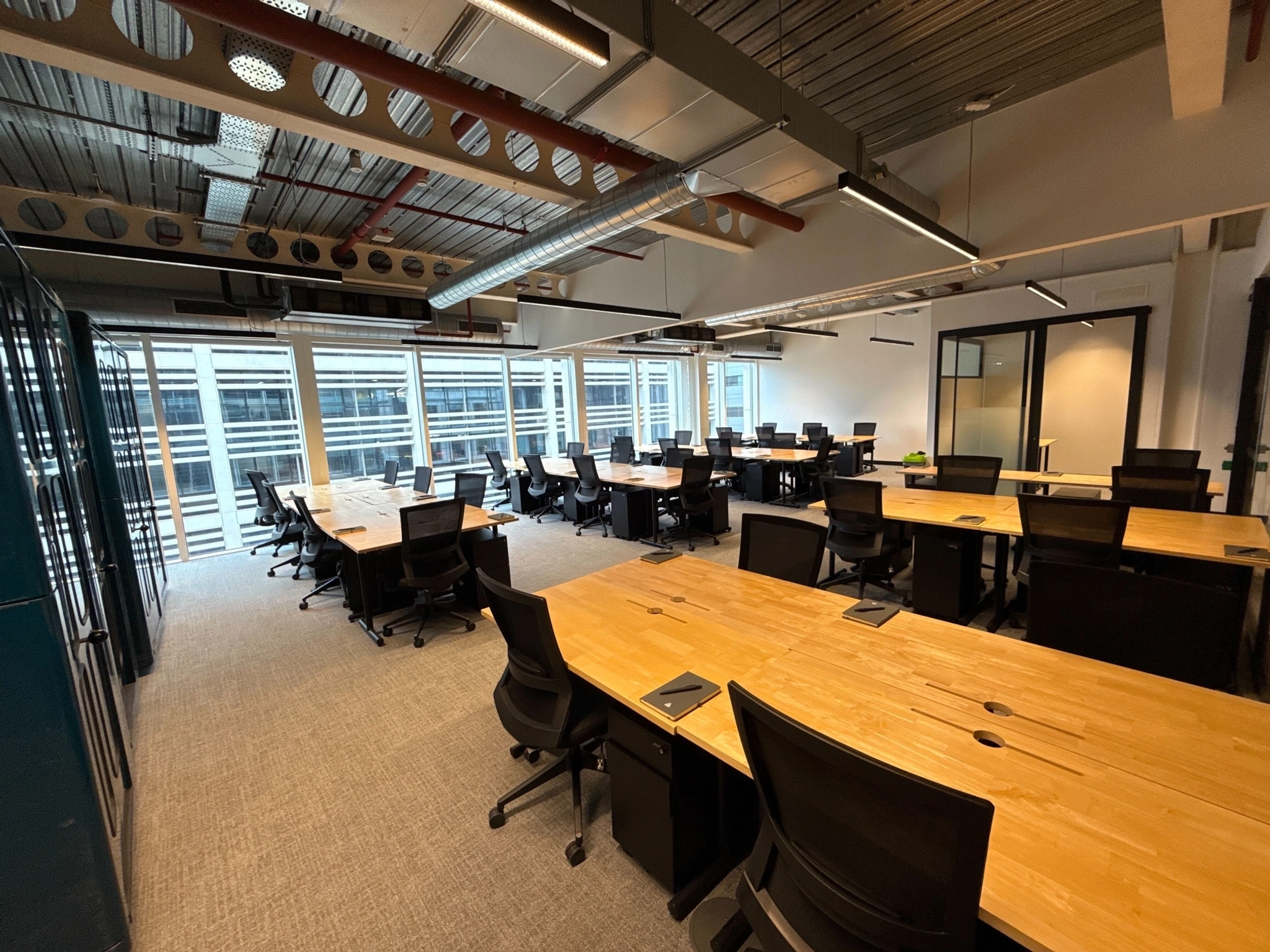 Modern open-plan workspace with wooden desks, black chairs, and large windows letting in natural light. Laptops and notepads are placed on some desks, creating an inviting coworking environment.