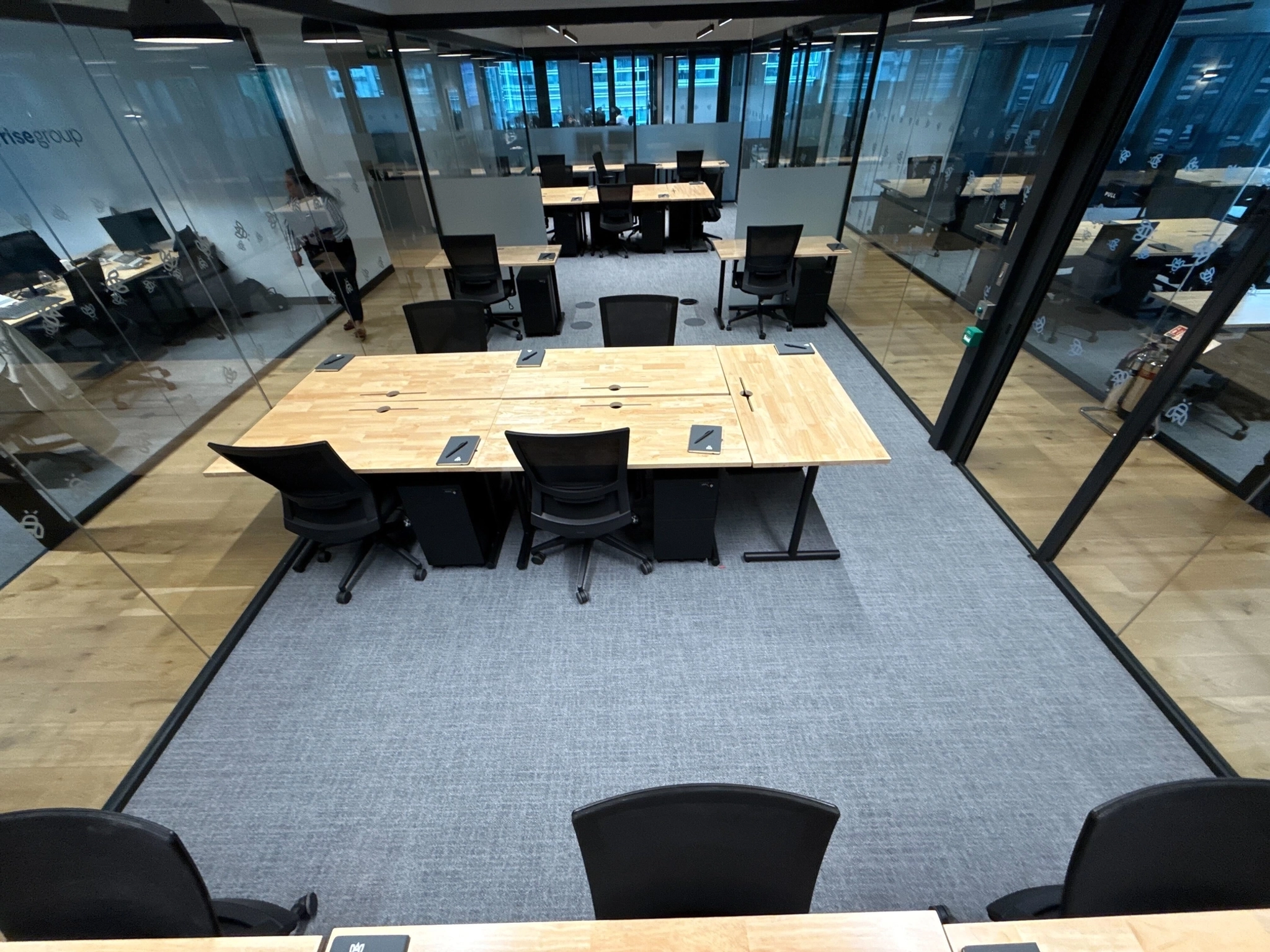 Modern coworking office space with wooden desks, black chairs, and glass walls, viewed from above; desks are arranged in rows with computers and chairs, and the room is brightly lit.