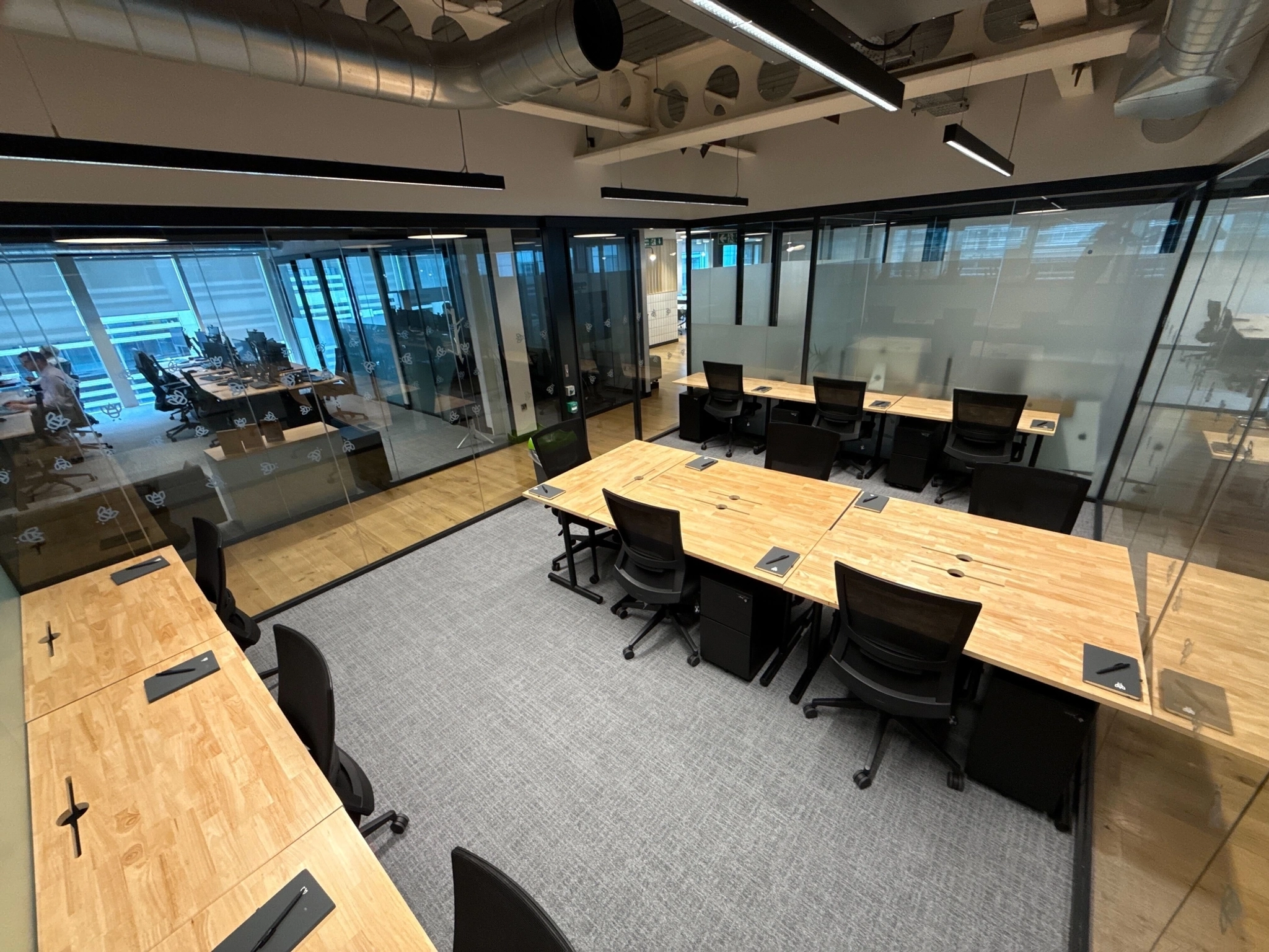 Moderne coworking kantoorruimte in Londen met houten bureaus, zwarte stoelen en glazen wanden; sommige werkruimtes zijn niet bezet en de aangrenzende ruimtes zijn zichtbaar door de glazen wanden.